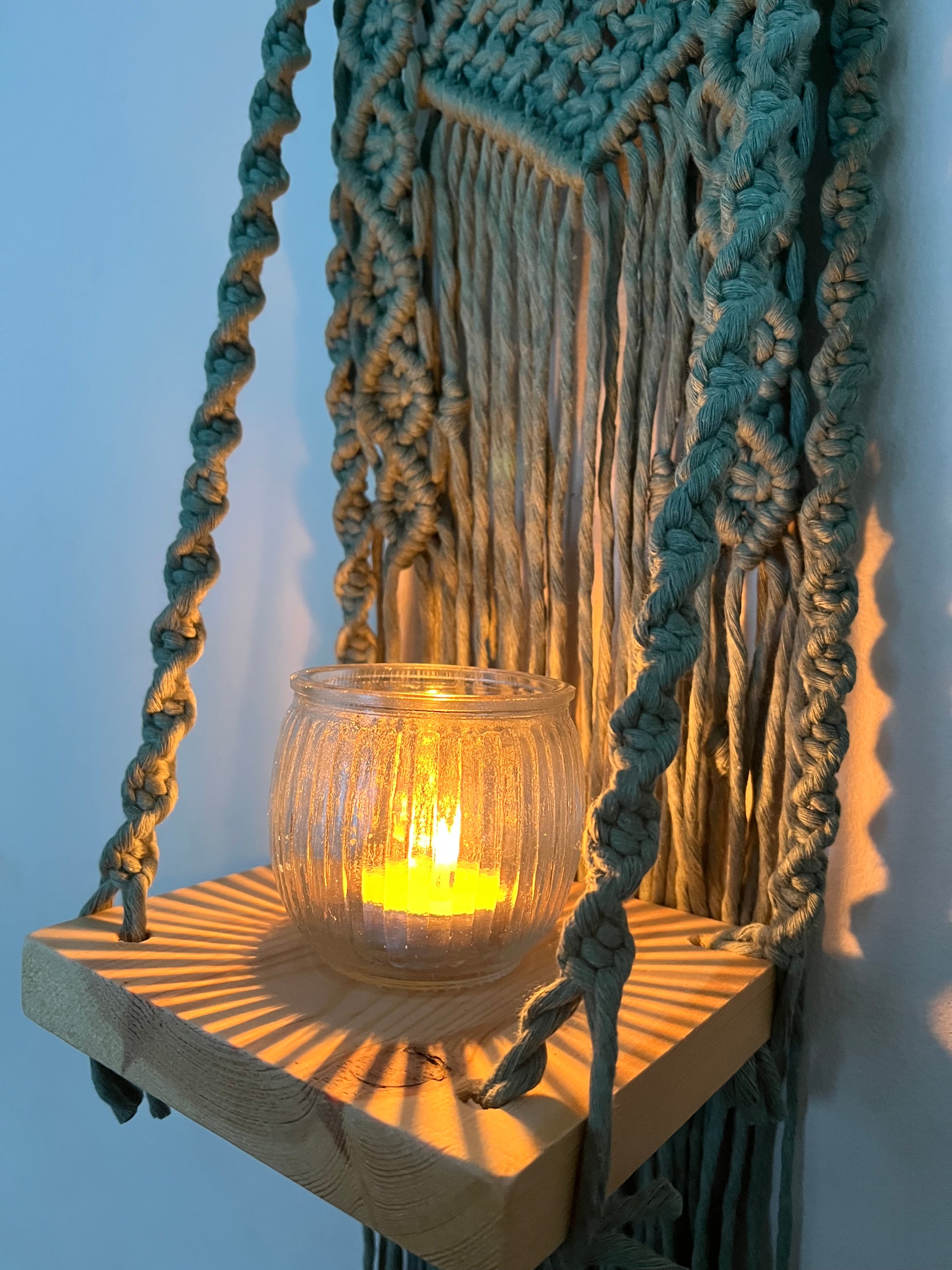 Handmade macrame boho style shelf, made with sage green recycled cotton on a solid wooden dowel, with a small square shelf. The shelf has an intricately knotted pattern, and displays a small lit candle in a glass jar. 