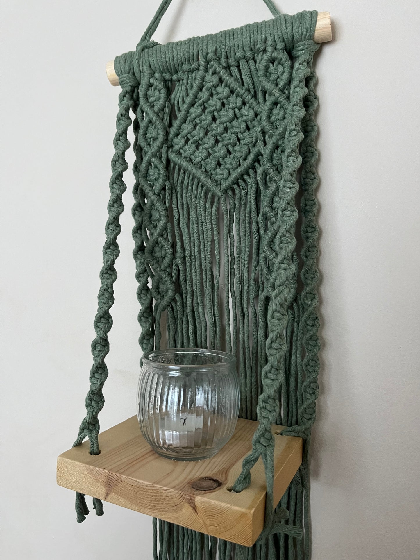 Handmade macrame boho style shelf, made with sage green recycled cotton on a solid wooden dowel, with a small square shelf. The shelf has an intricately knotted pattern, and displays a glass candle jar. 