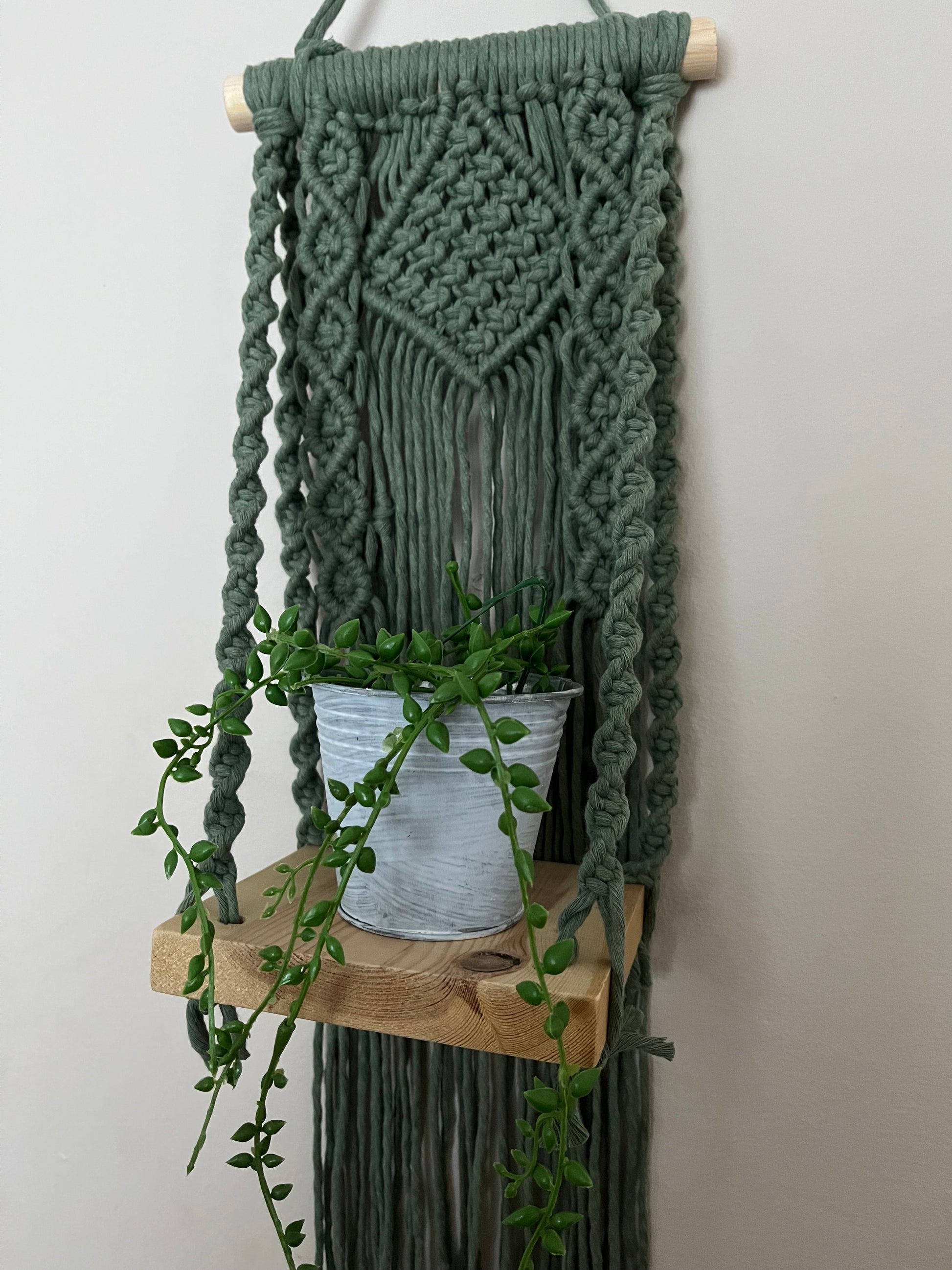 Handmade macrame boho style shelf, made with sage green recycled cotton on a solid wooden dowel, with a small square shelf. The shelf has an intricately knotted pattern, and displays a small artificial plant. 