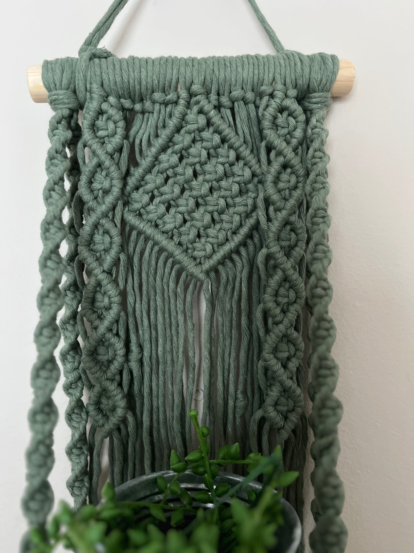 Handmade macrame boho style shelf, made with sage green recycled cotton on a solid wooden dowel, with a small square shelf. The shelf has an intricately knotted pattern.