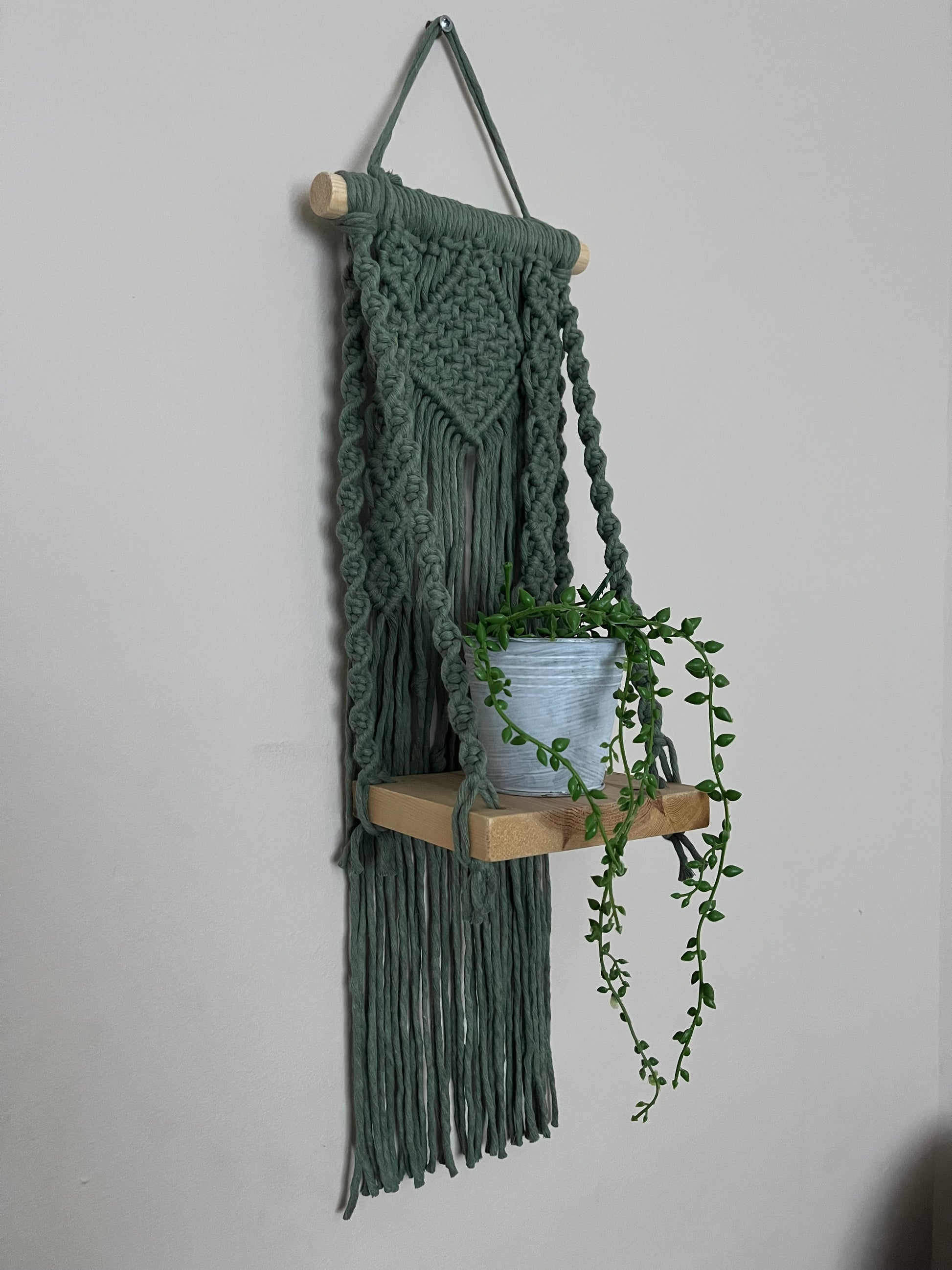 Handmade macrame boho style shelf, made with sage green recycled cotton on a solid wooden dowel, with a small square shelf. The shelf has an intricately knotted pattern, and displays a small artificial plant. 