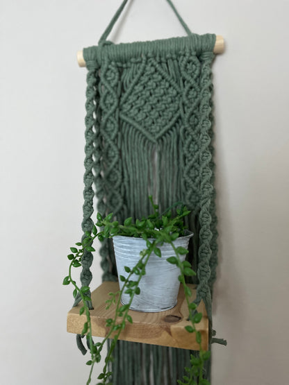 Handmade macrame boho style shelf, made with sage green recycled cotton on a solid wooden dowel, with a small square shelf. The shelf has an intricately knotted pattern, and displays a small artificial plant. 