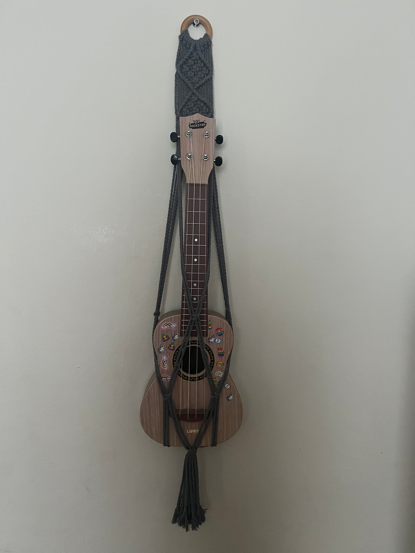 Soprano sized ukulele, with stickers on it, hanging against a plain wall in a dark grey macrame hanger. 