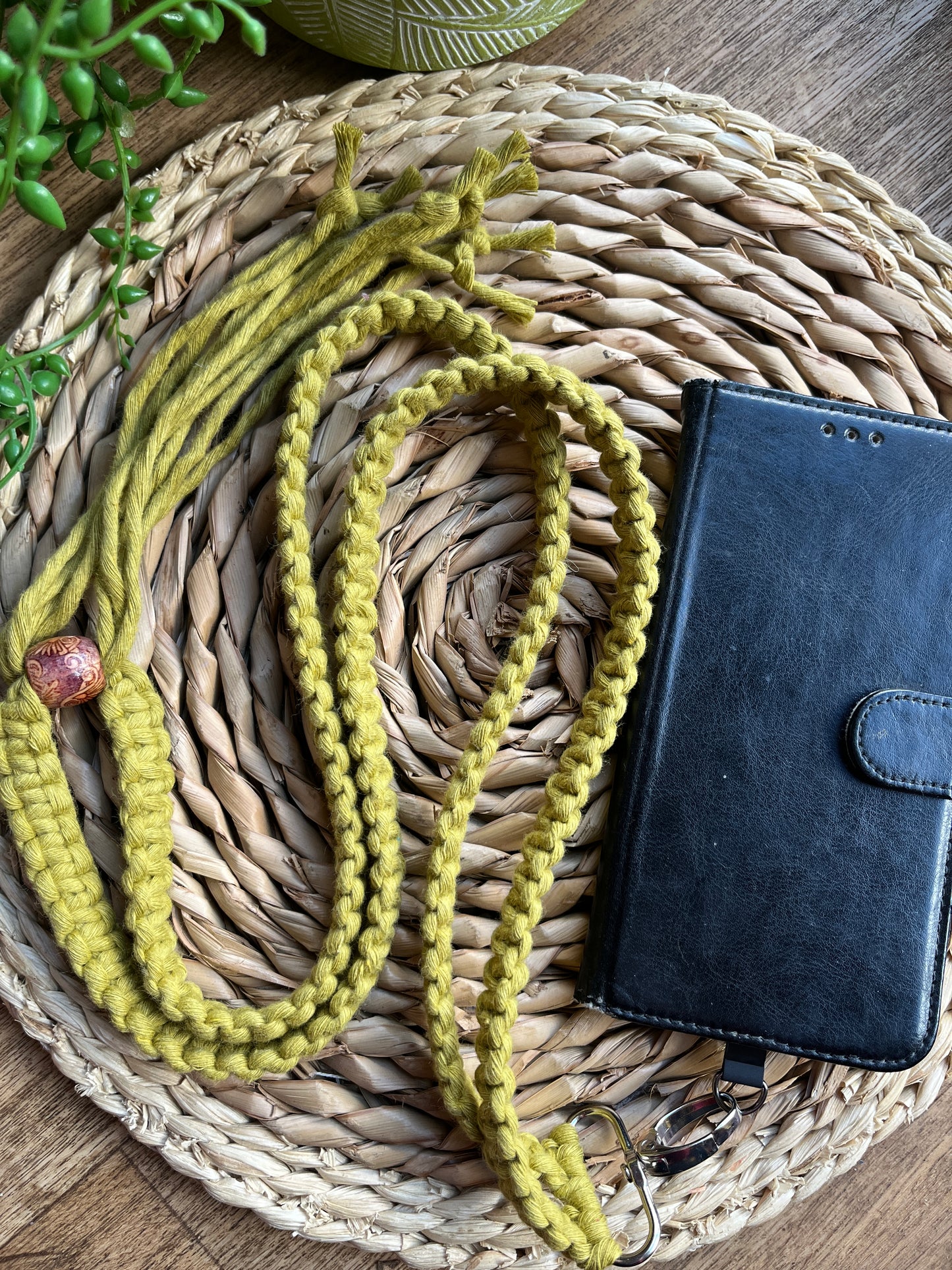 A long crossbody phone strap made with green yarn, a patterned wooden bead and metal clasp attached to a black leather phone case on a round wicker placemat. 