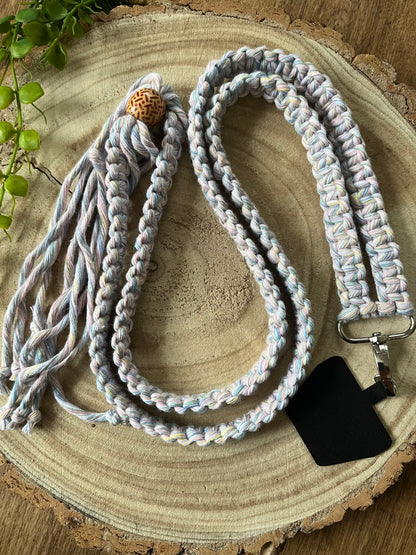 A long crossbody phone strap made with pastel yarn, a patterned wooden bead and metal clasp has a universal tether attached to it, and is coiled on a round wooden surface.
