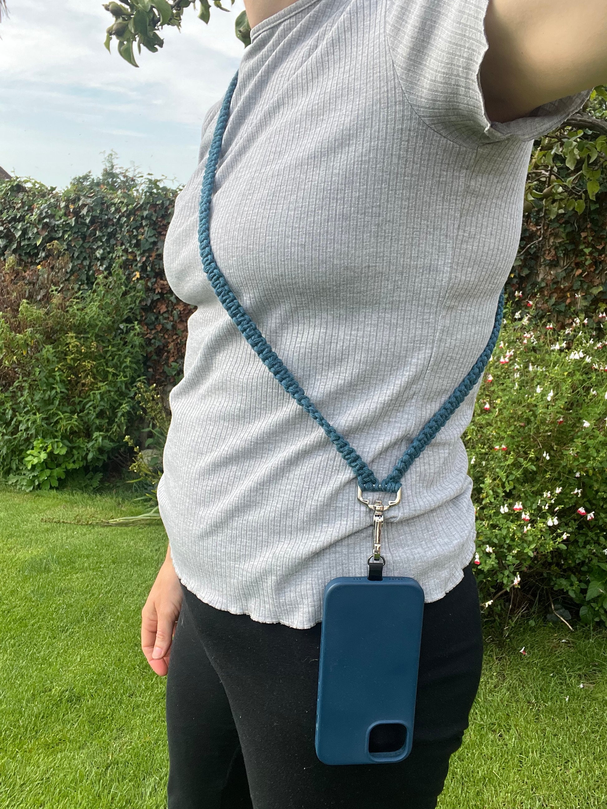 A person wearing a light grey ribbed short-sleeve shirt and black leggings holds a blue phone case on a long, crossbody phone strap made with petrol blue yarn. 