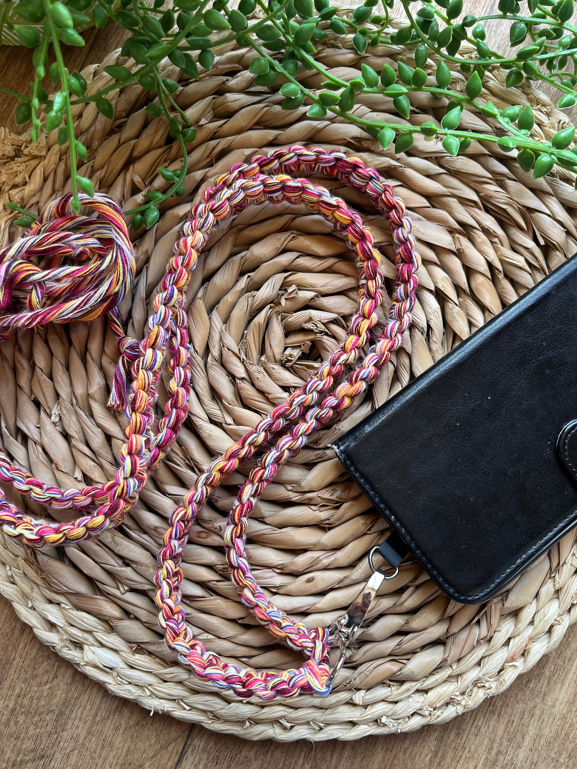 A long crossbody phone strap made with multicoloured yarn, attached to a black leather phone case on a round wicker placemat. 