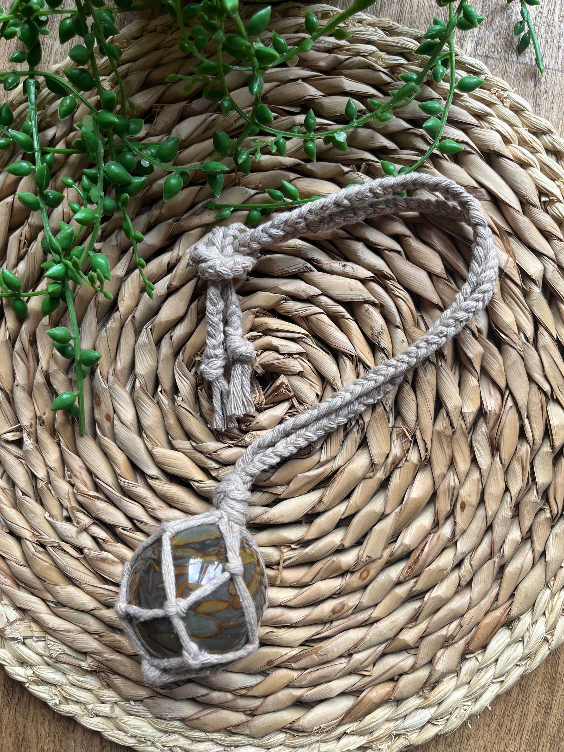 A large bamboo jasper sphere held neatly inside a knotted macrame hanger made with taupe recycled cotton, laid across a round woven mat with some green plant vines. 