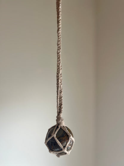 A large bamboo jasper sphere held neatly inside a knotted macrame hanger made with taupe recycled cotton, hanging in front of a plain background. 