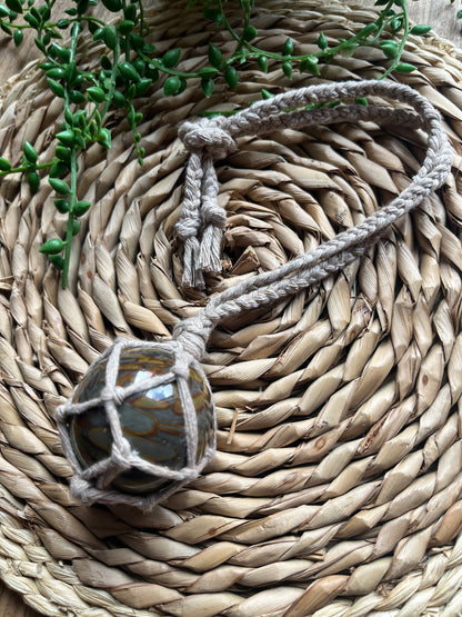 Close up of a large bamboo jasper sphere held neatly inside a knotted macrame hanger made with taupe recycled cotton, laid across a round woven mat with some green plant vines. 