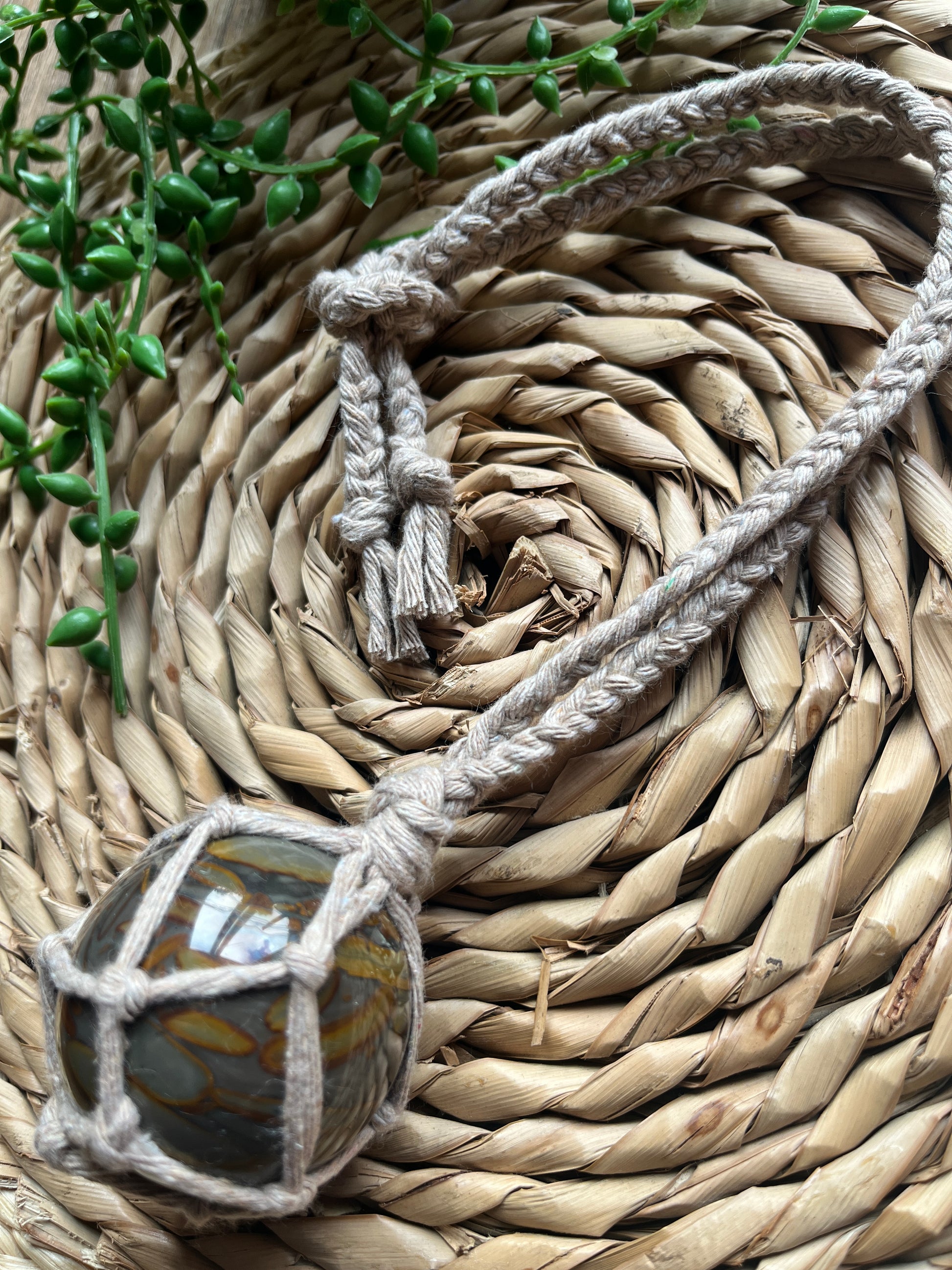 A large bamboo jasper sphere held neatly inside a knotted macrame hanger made with taupe recycled cotton, laid across a round woven mat with some green plant vines. 