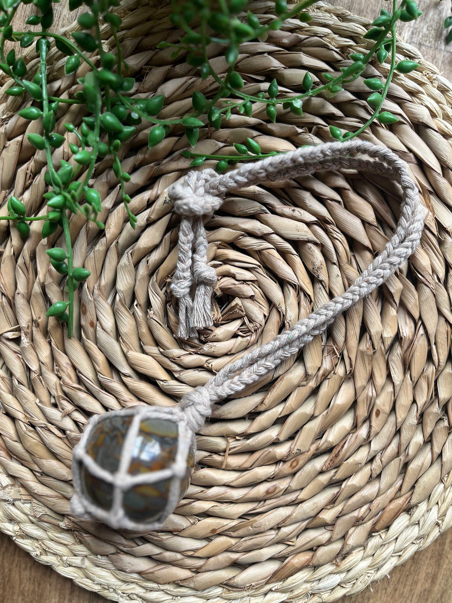 A large bamboo jasper sphere held neatly inside a knotted macrame hanger made with taupe recycled cotton, laid across a round woven mat with some green plant vines. 
