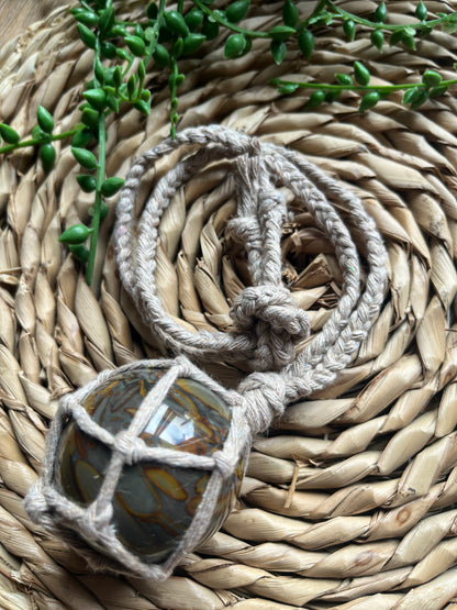 A large bamboo jasper sphere held neatly inside a knotted macrame hanger made with taupe recycled cotton, coiled on a round woven mat with some green plant vines. 