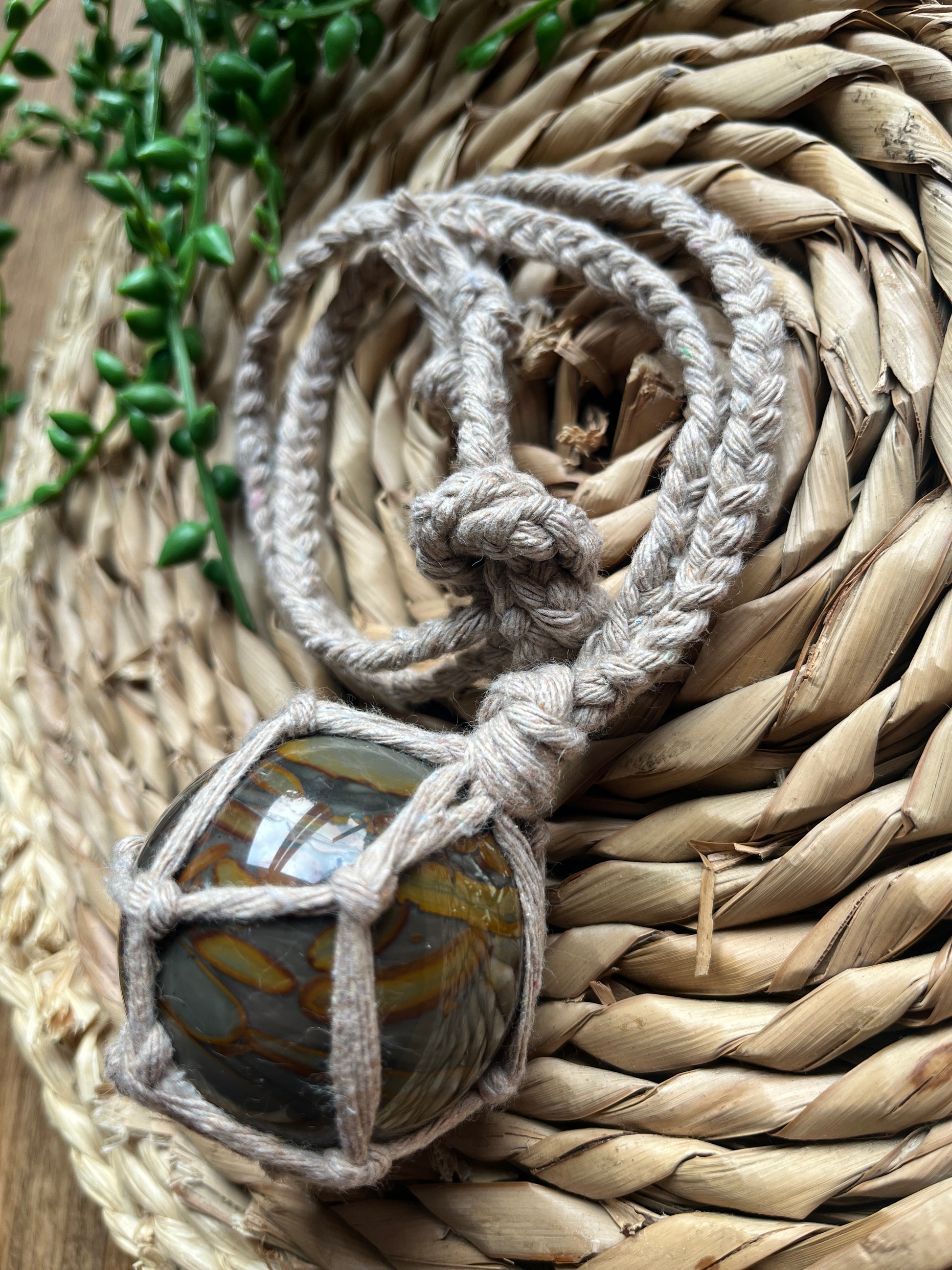 Close up of a large bamboo jasper sphere held neatly inside a knotted macrame hanger made with taupe recycled cotton, coiled on a round woven mat with some green plant vines. 