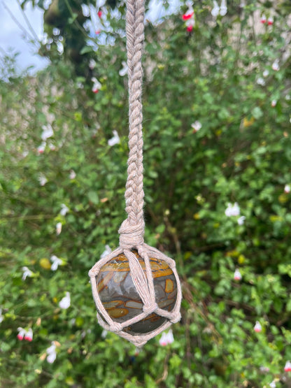 A large bamboo jasper sphere held neatly inside a knotted macrame hanger made with taupe recycled cotton, hanging outside in front of lush green plants. 