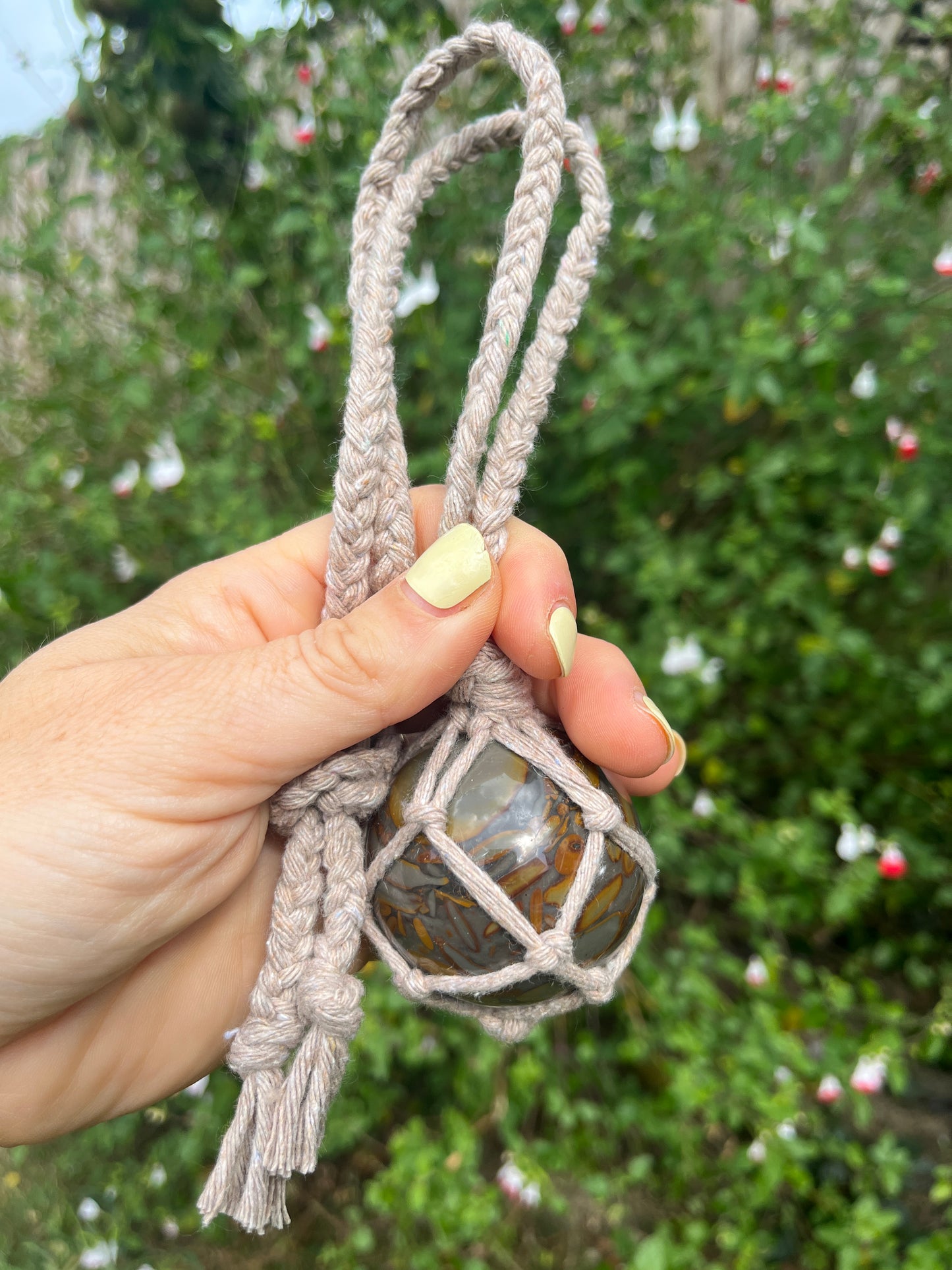 A hand holding a large bamboo jasper sphere held neatly inside a knotted macrame hanger made with taupe recycled cotton, outside with lush green plants in the background. 