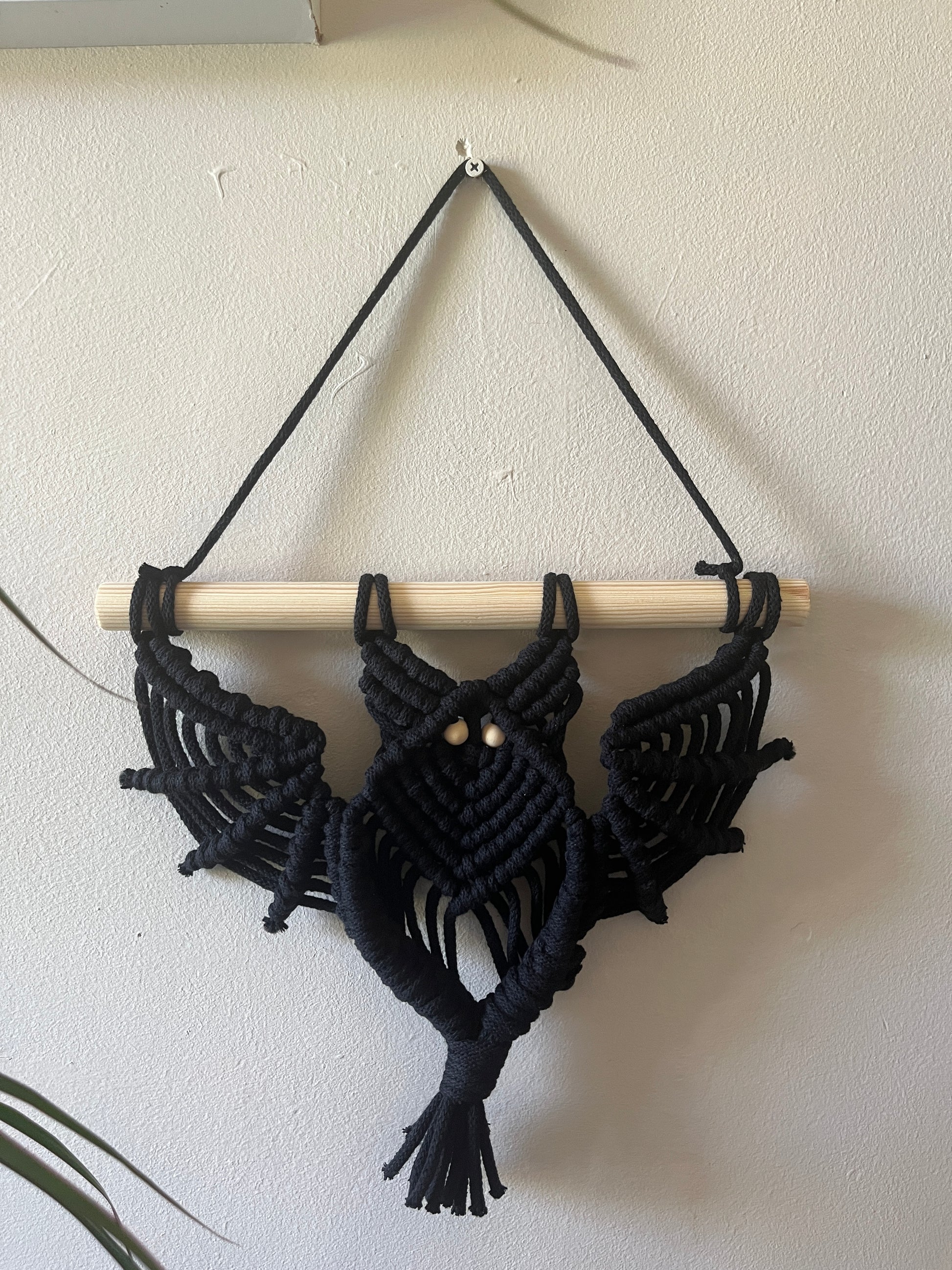 Black macrame bat wall hanging with wooden bead eyes on a wooden dowel, hanging against a beige painted wall