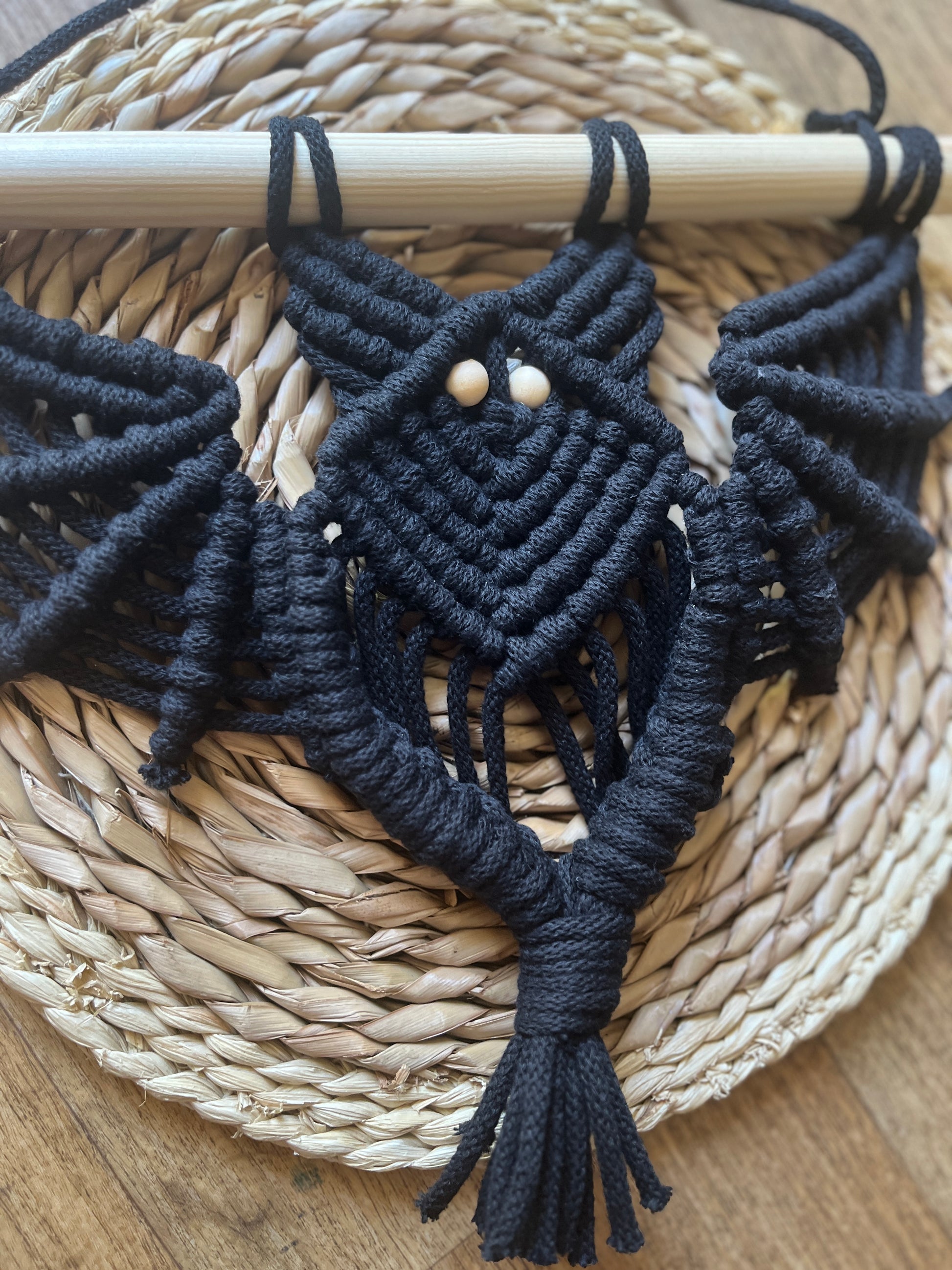 Close up of a black macrame bat wall hanging with wooden bead eyes on a wooden dowel, on a wicker surface.
