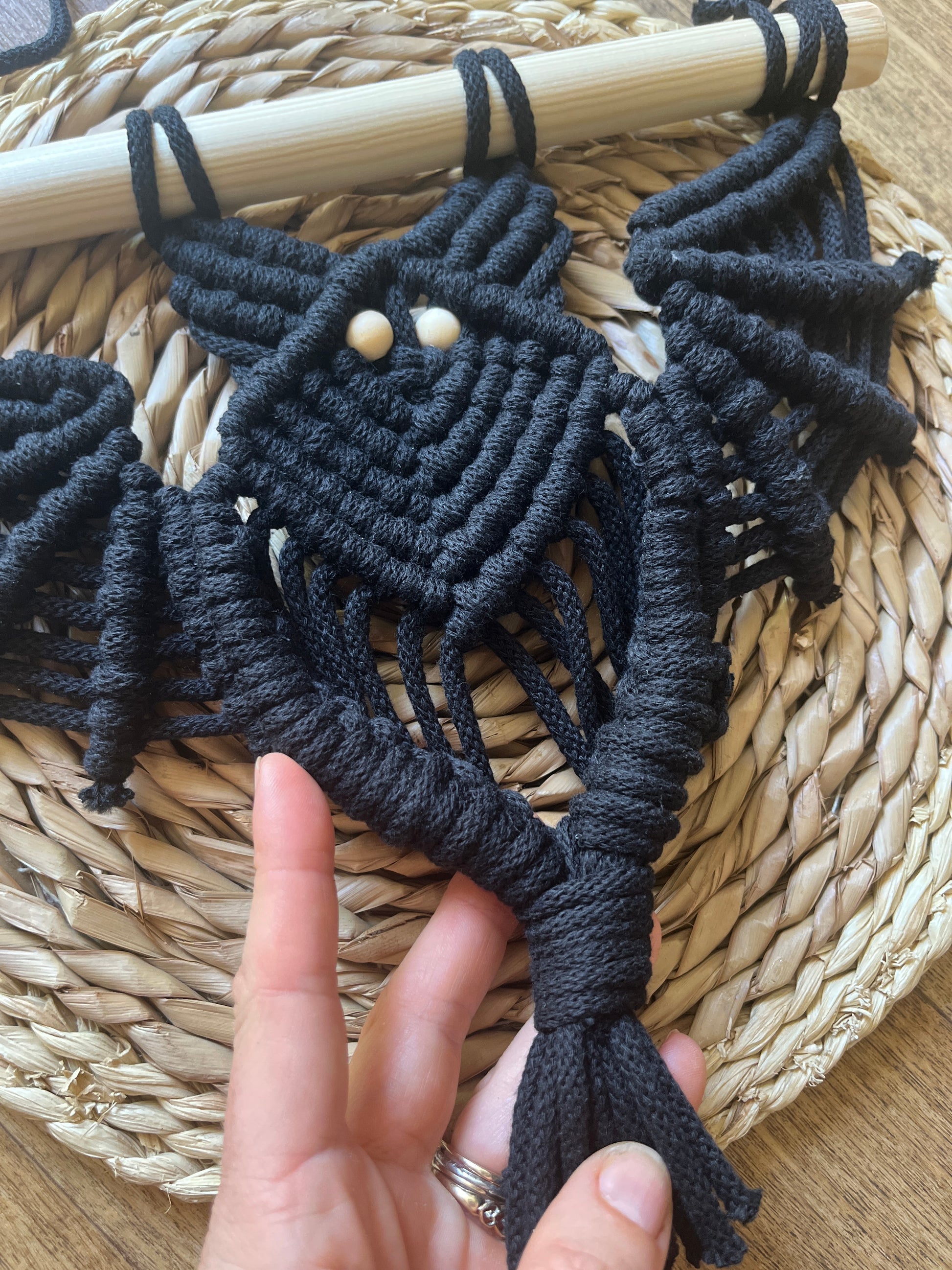A hand holding the tail of a black macrame bat wall hanging with wooden bead eyes on a wooden dowel, on a wicker mat surface. 