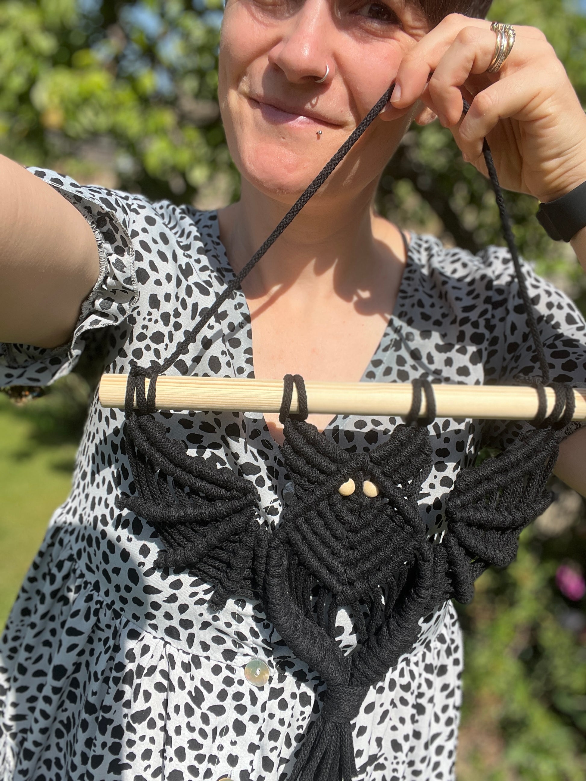 A person stands outside wearing a black and white spotted dress and smiling towards the camera, she is holding a macrame bat wall hanging 