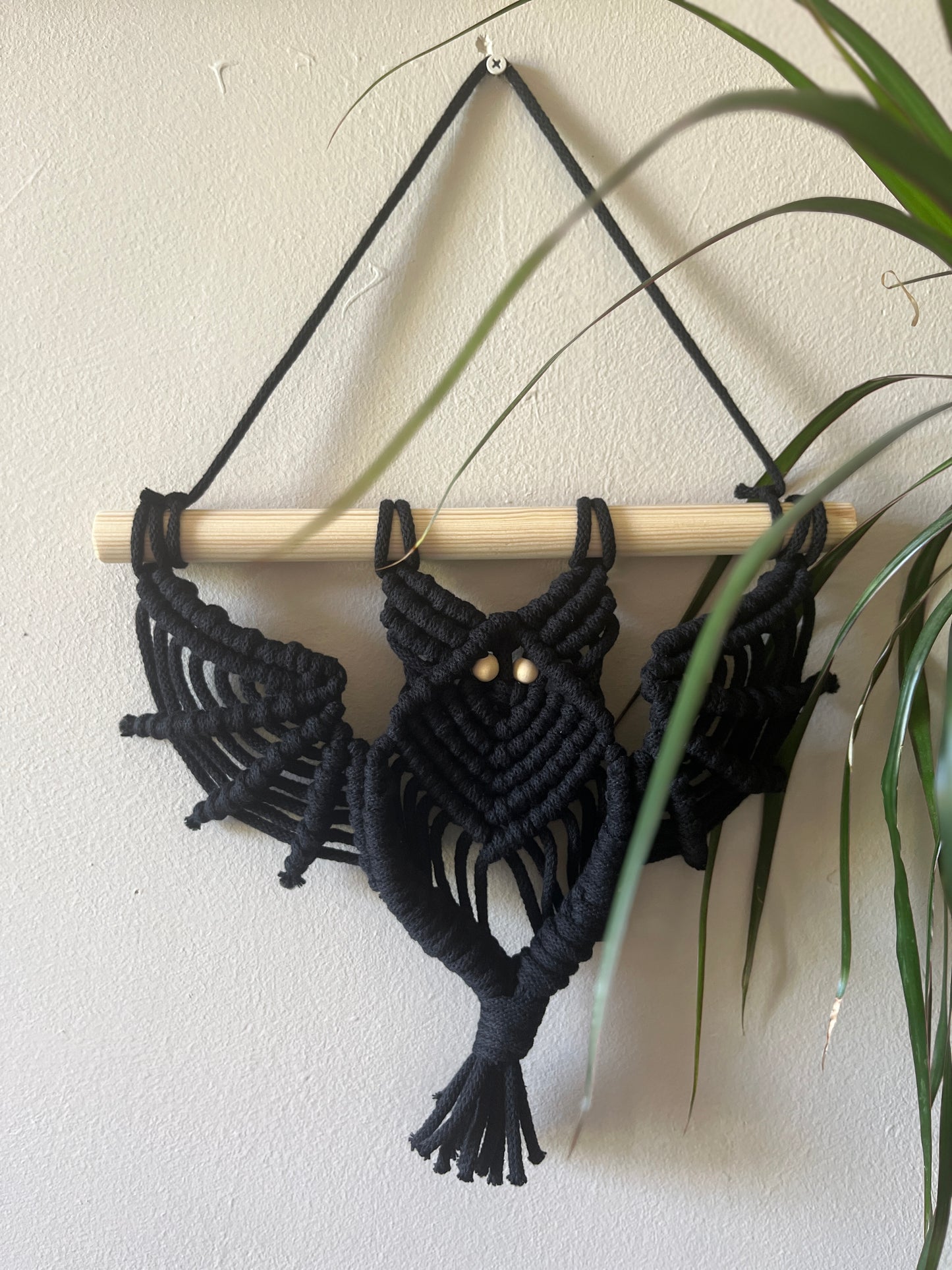 Black macrame wall hanging in the shape of a bat, with a wooden dowel on a plain wall.