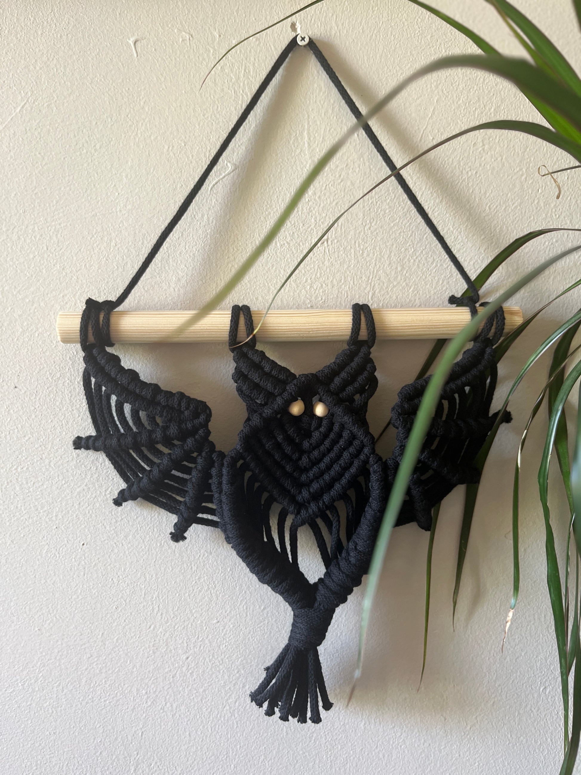 Black macrame wall hanging in the shape of a bat, with a wooden dowel on a plain wall.