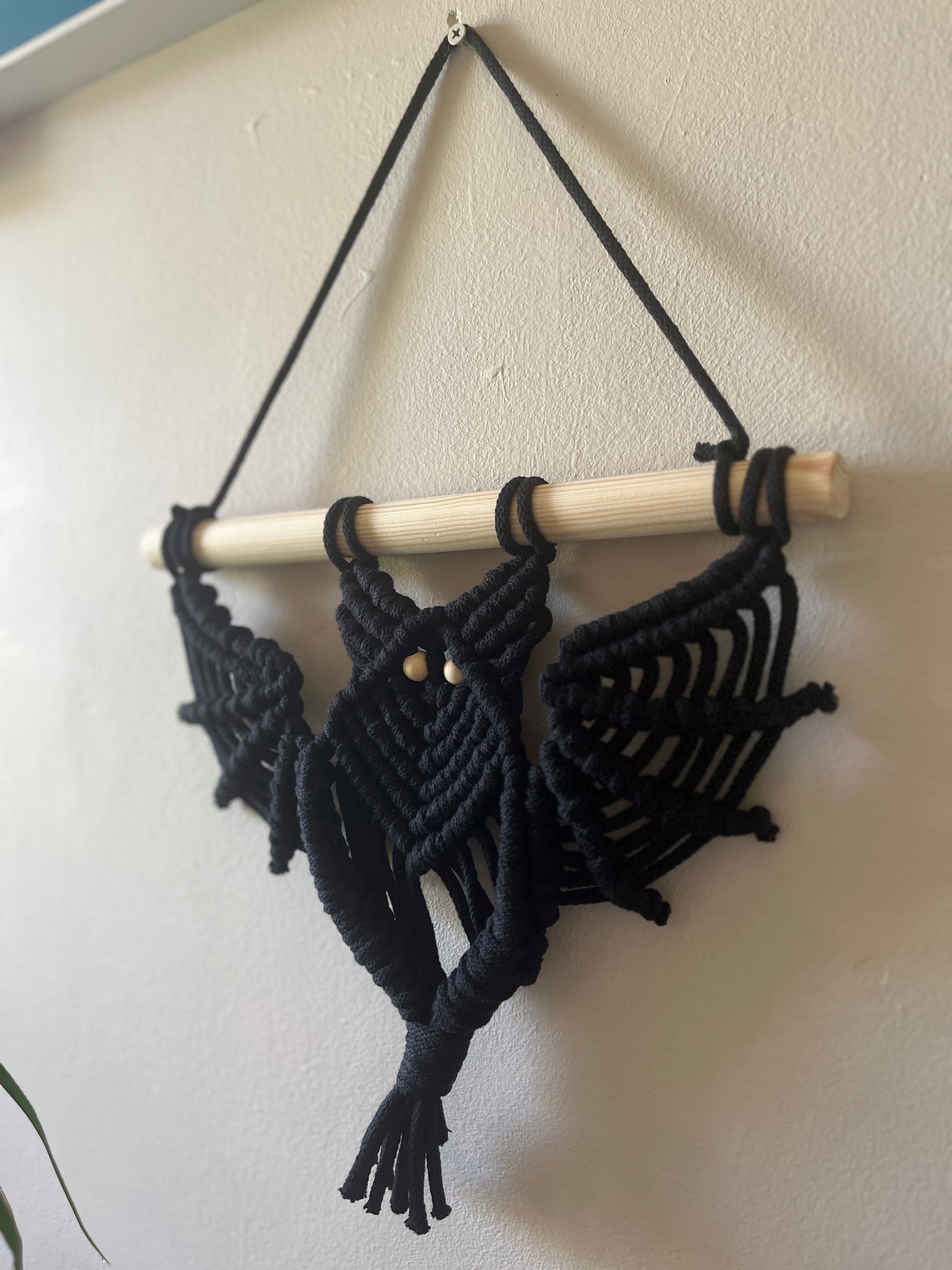 Black macrame bat shaped wall hanging with wooden bead eyes on a wooden dowel, hanging against a beige painted wall