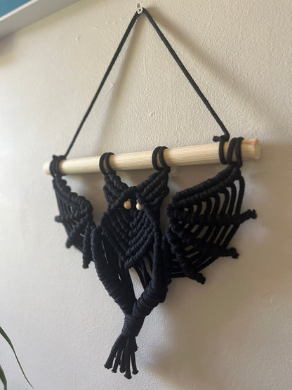 Black macrame bat shaped wall hanging with wooden bead eyes on a wooden dowel, hanging against a beige painted wall
