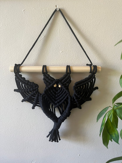 Black macrame bat shaped wall hanging with wooden bead eyes on a wooden dowel, hanging against a beige painted wall, with. green houseplant just visible to the right.