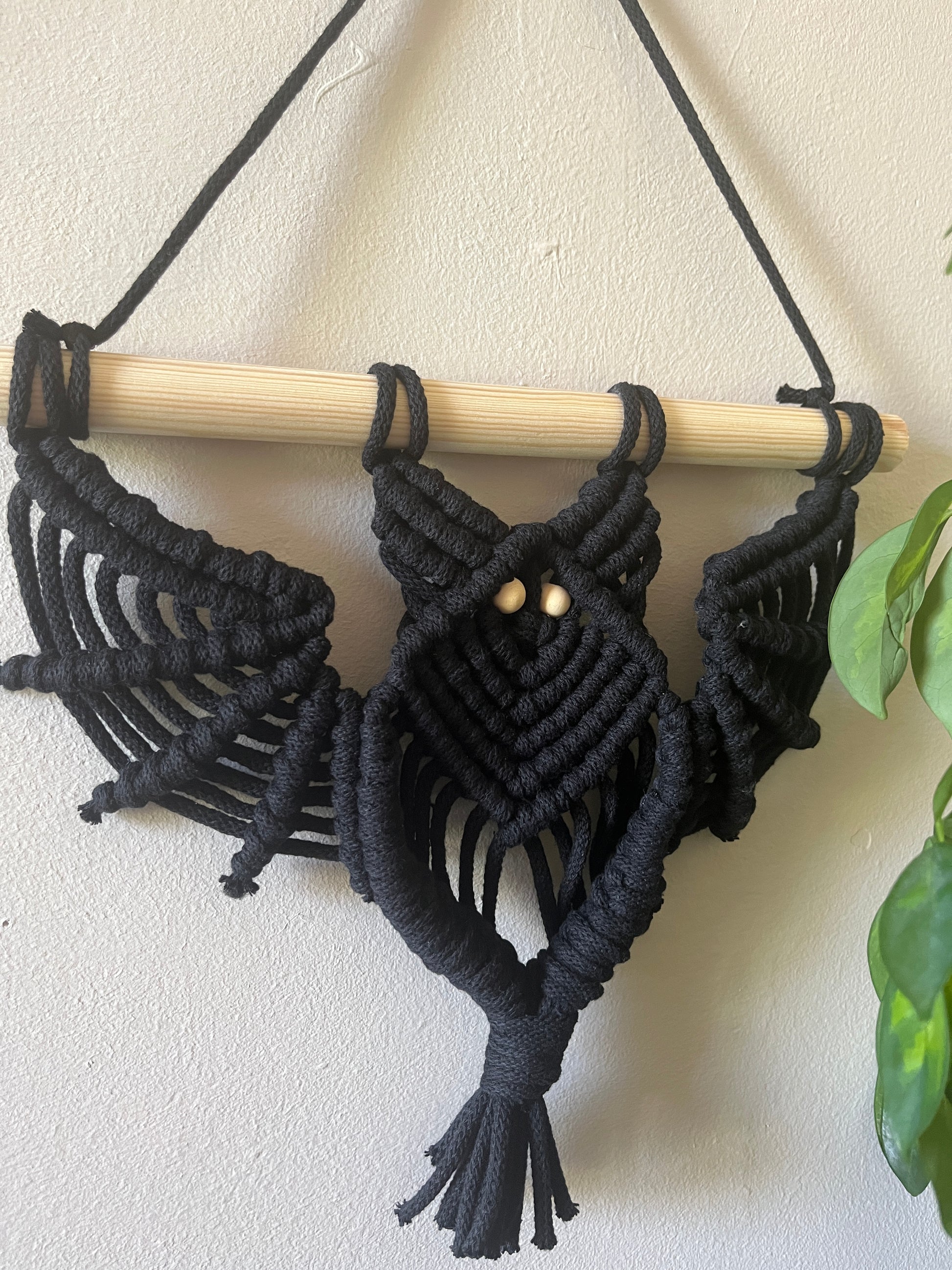 Close up of a black macrame bat wall hanging with wooden bead eyes on a wooden dowel, hanging against a beige painted wall