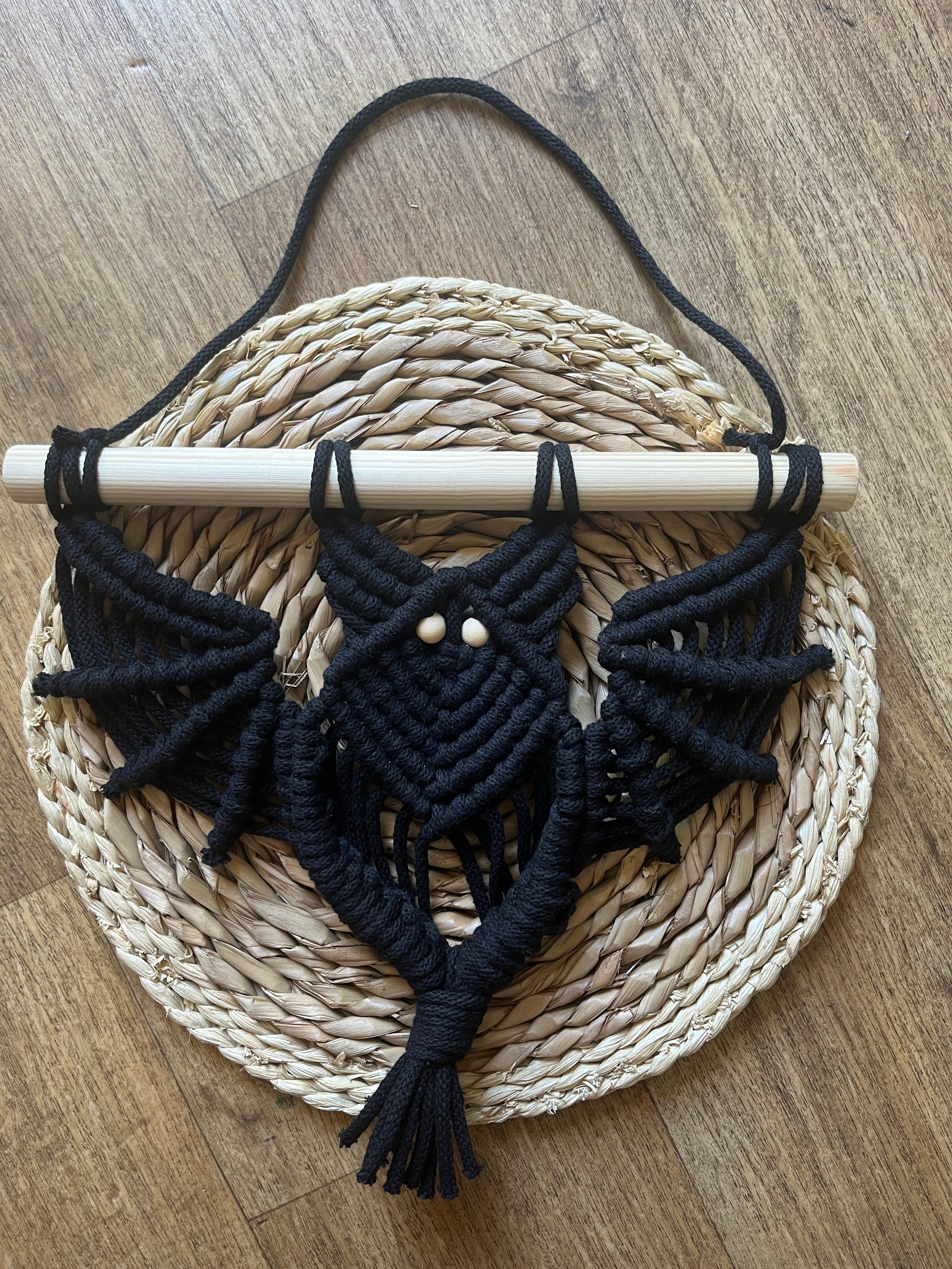 Black macrame bat shaped wall hanging with wooden bead eyes on a wooden dowel, laid flat across a round wicker mat. 
