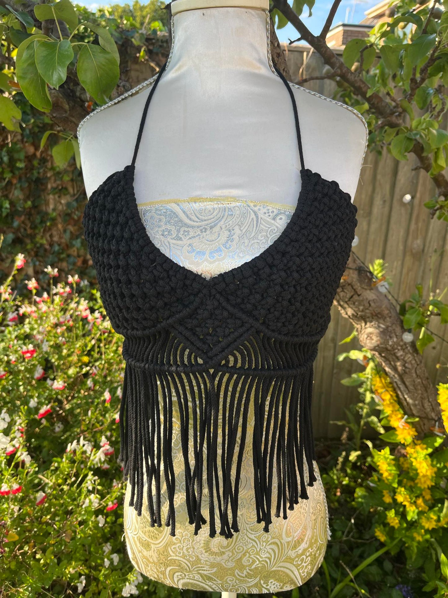 Black macrame halter top with fringe on a mannequin against a garden background