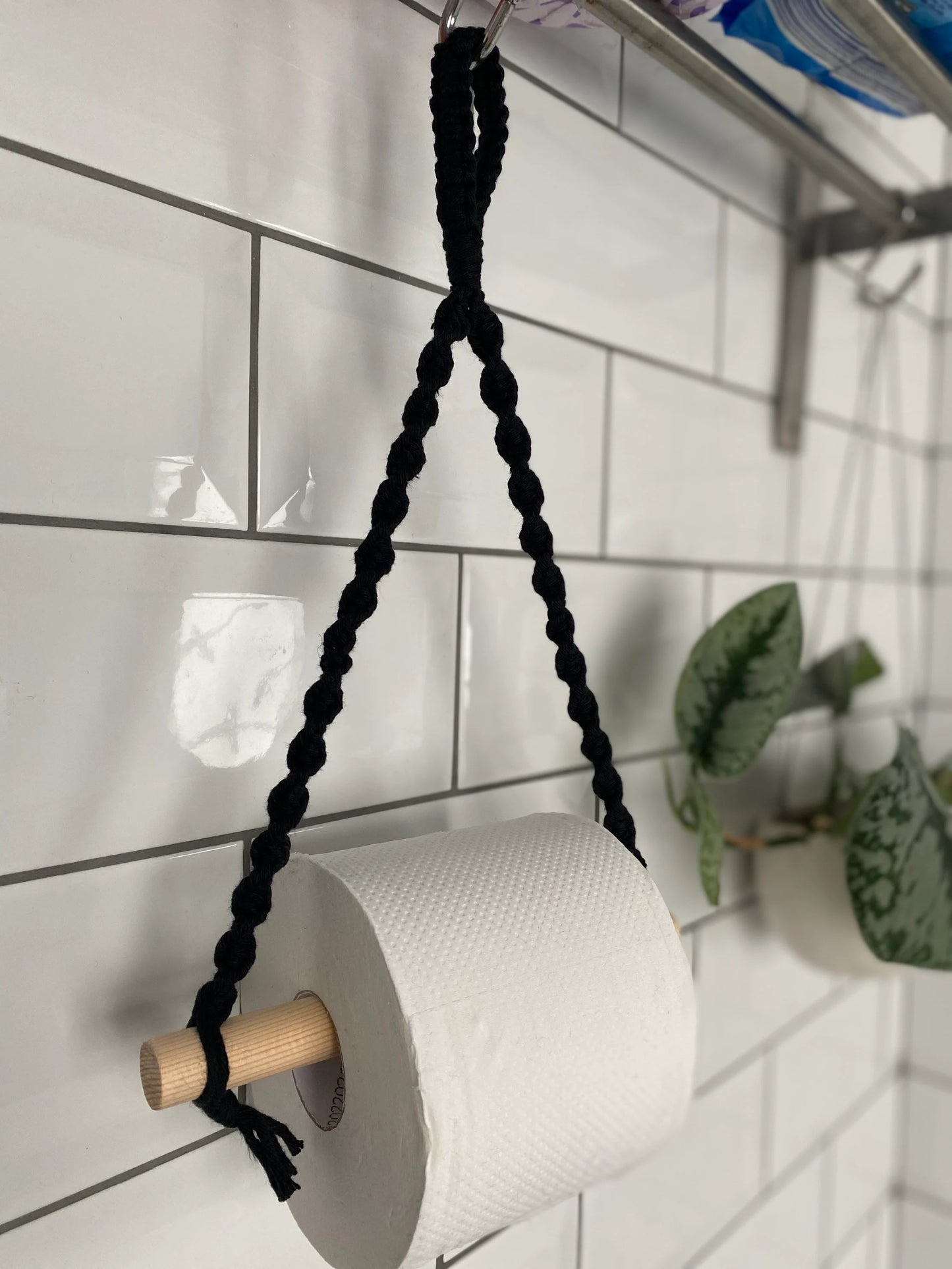 A Macra-Made-With-Love Single toilet roll holder hangs from a black, braided cord attached to a wooden dowel. The bathroom accessory is mounted on a white tiled wall. A small plant with green leaves is placed in the background on a hanging shelf, adding eco-friendly decor to the space.