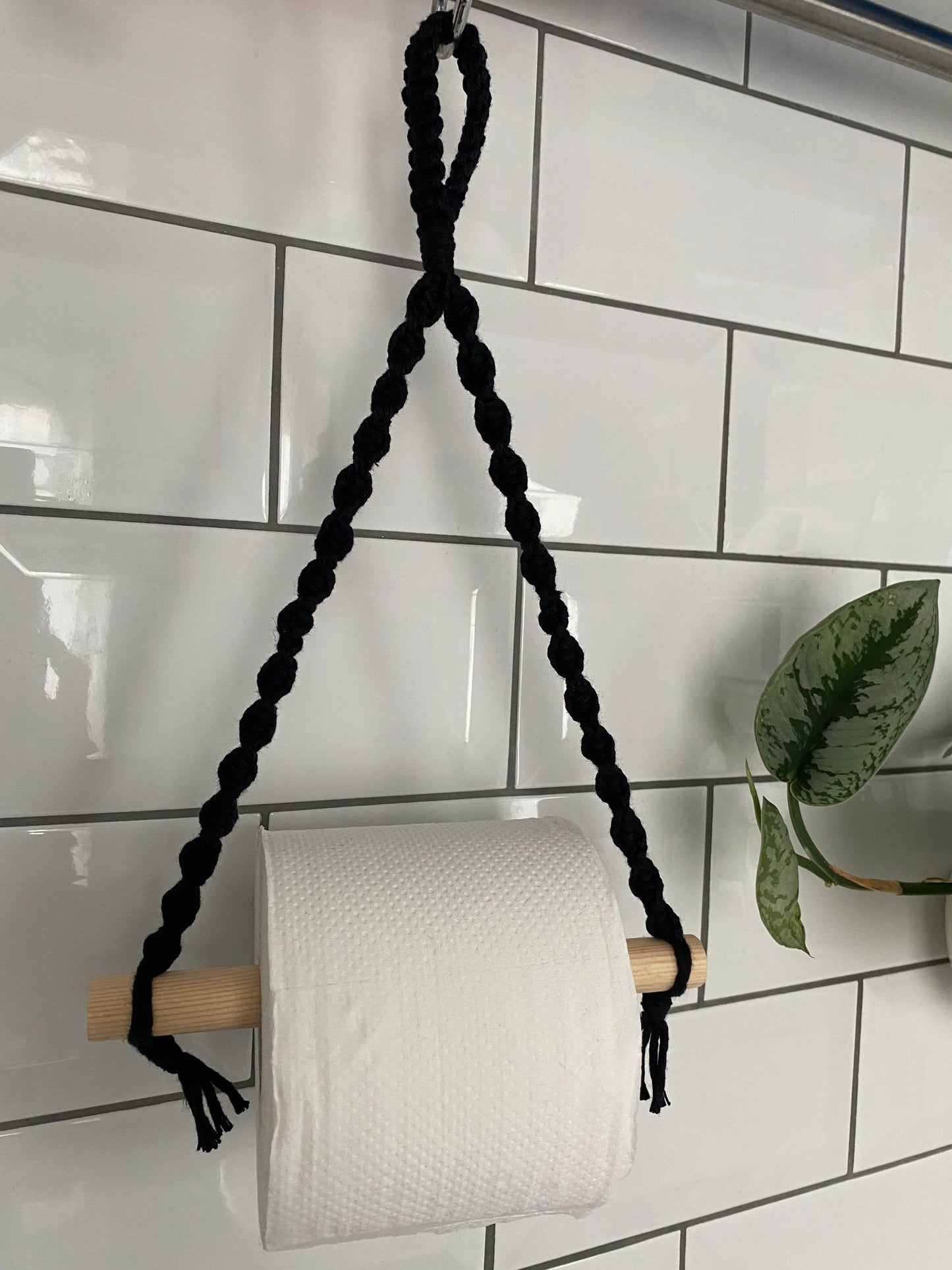 A handmade Macra-Made-With-Love Single toilet roll holder hangs on a wooden dowel supported by a black braided cord, adding eco-friendly decor to your bathroom. It's set against a white tiled wall with a green leafy plant partially visible on the right side.