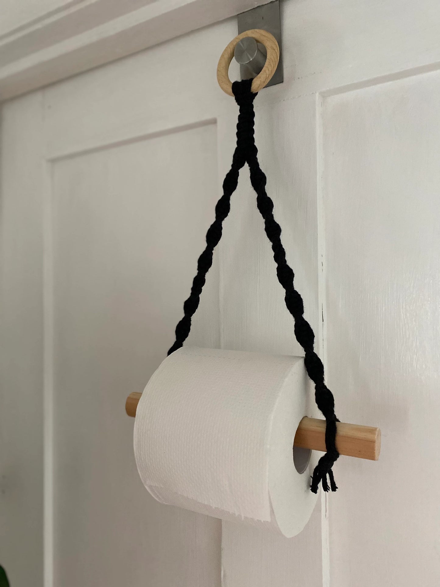 A Macra-Made-With-Love Single toilet roll holder hangs on a handmade macrame toilet roll holder, crafted from natural wood and braided rope. This eco-friendly bathroom accessory is suspended by black rope from a hook mounted on a white wooden door, adding a simple and rustic charm.