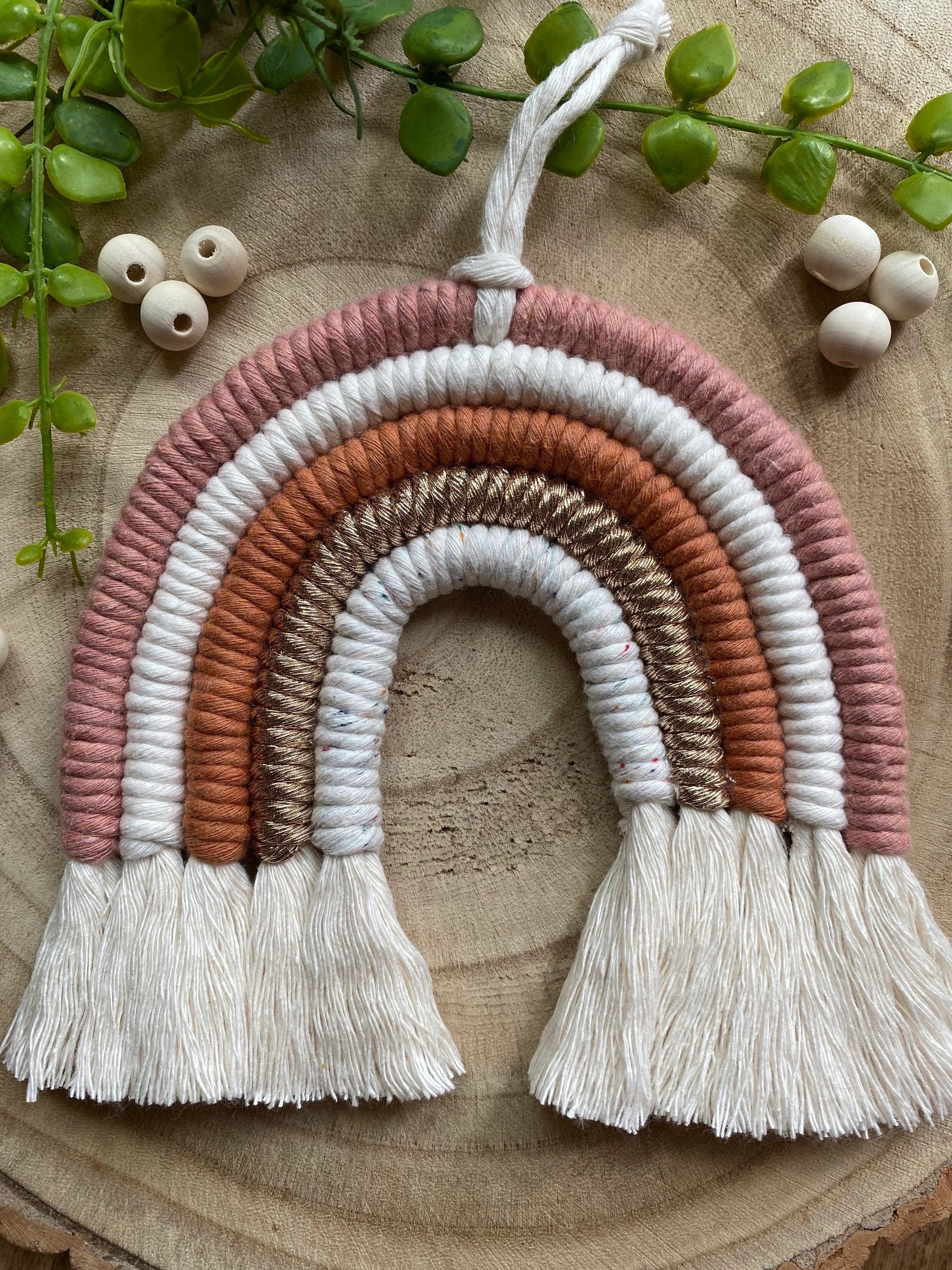 A handmade macrame rainbow wall hanging in shades of pink, white, and gold, with tassels on the ends. Displayed on a wooden board with green plant vines and wooden beads. 