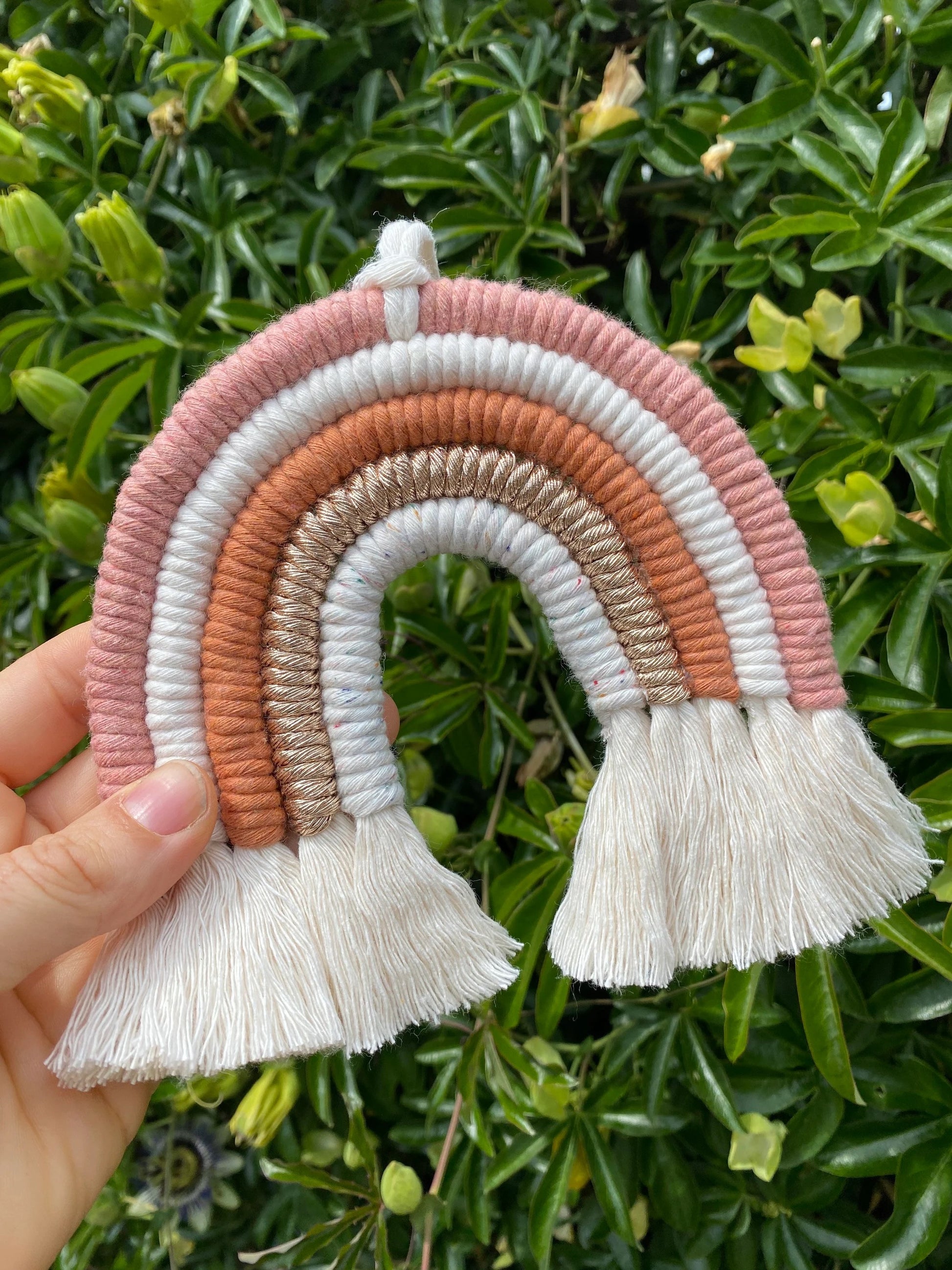 A hand holding a handmade macrame rainbow wall hanging in shades of pink, white, and gold, with tassels on the ends in front of lush green plants, outside. 