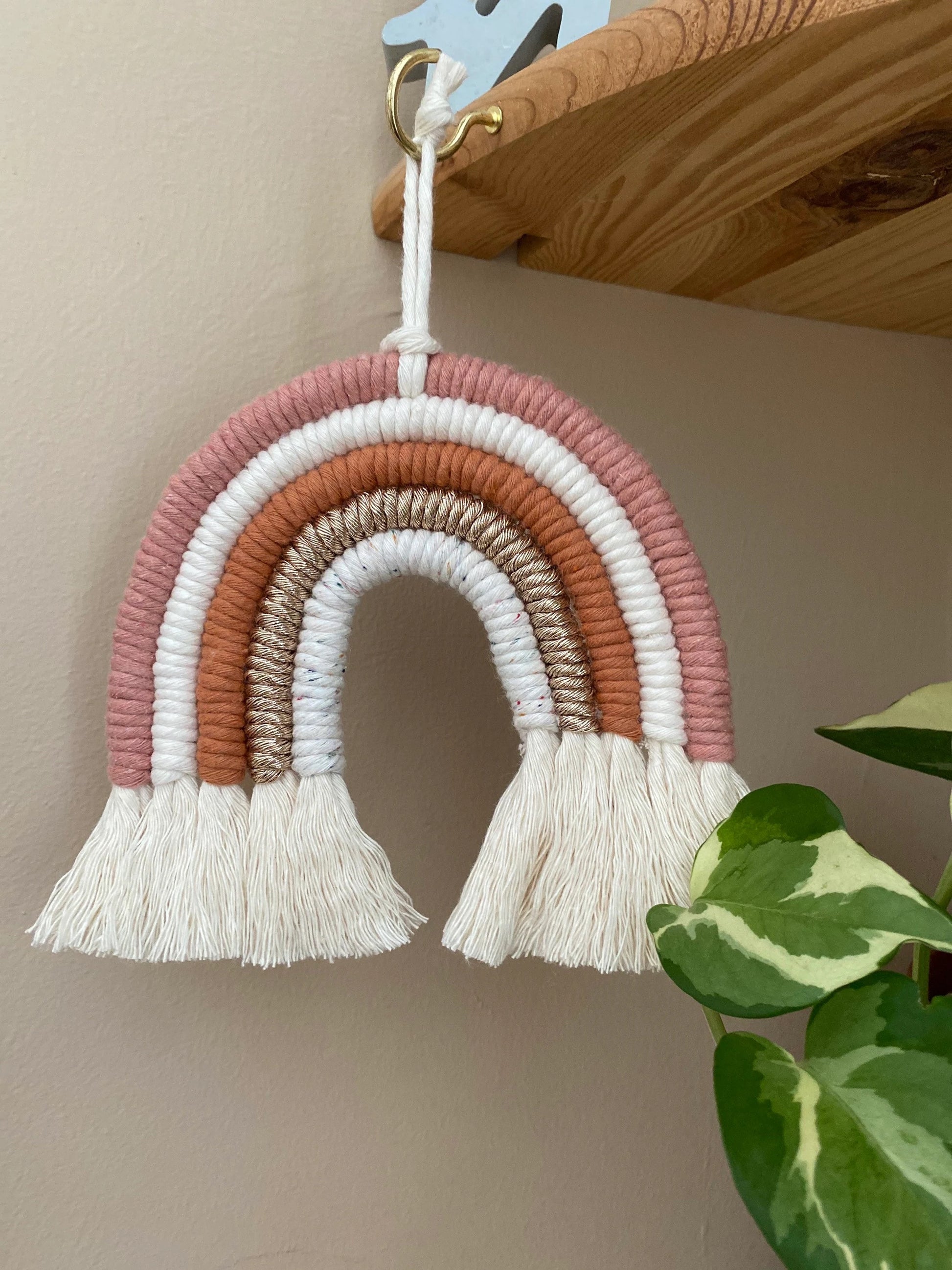 A handmade macrame rainbow wall hanging in shades of pink, white, and gold, with tassels on the ends hanging from a wooden corner shelf. 