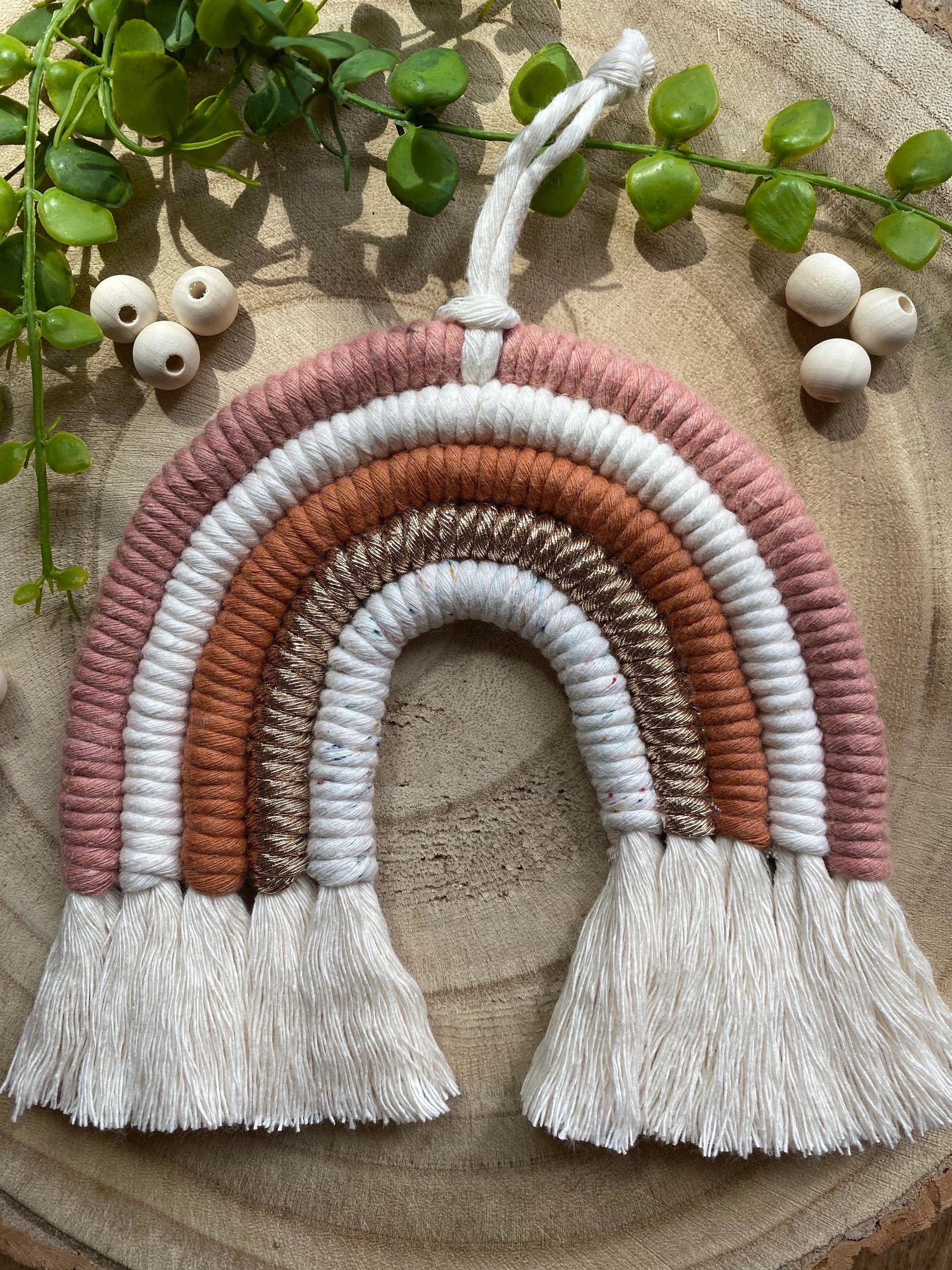 A handmade macrame rainbow wall hanging in shades of pink, white, and gold, with tassels on the ends. Displayed on a wooden board with green plant vines and wooden beads. 