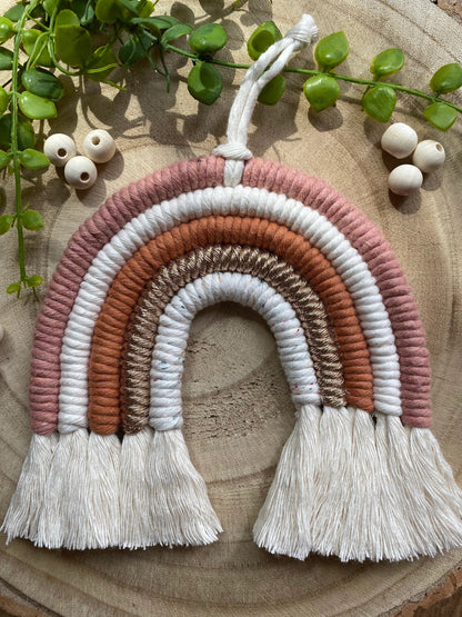 A handmade macrame rainbow wall hanging in shades of pink, white, and gold, with tassels on the ends. Displayed on a wooden board with green plant vines and wooden beads. 