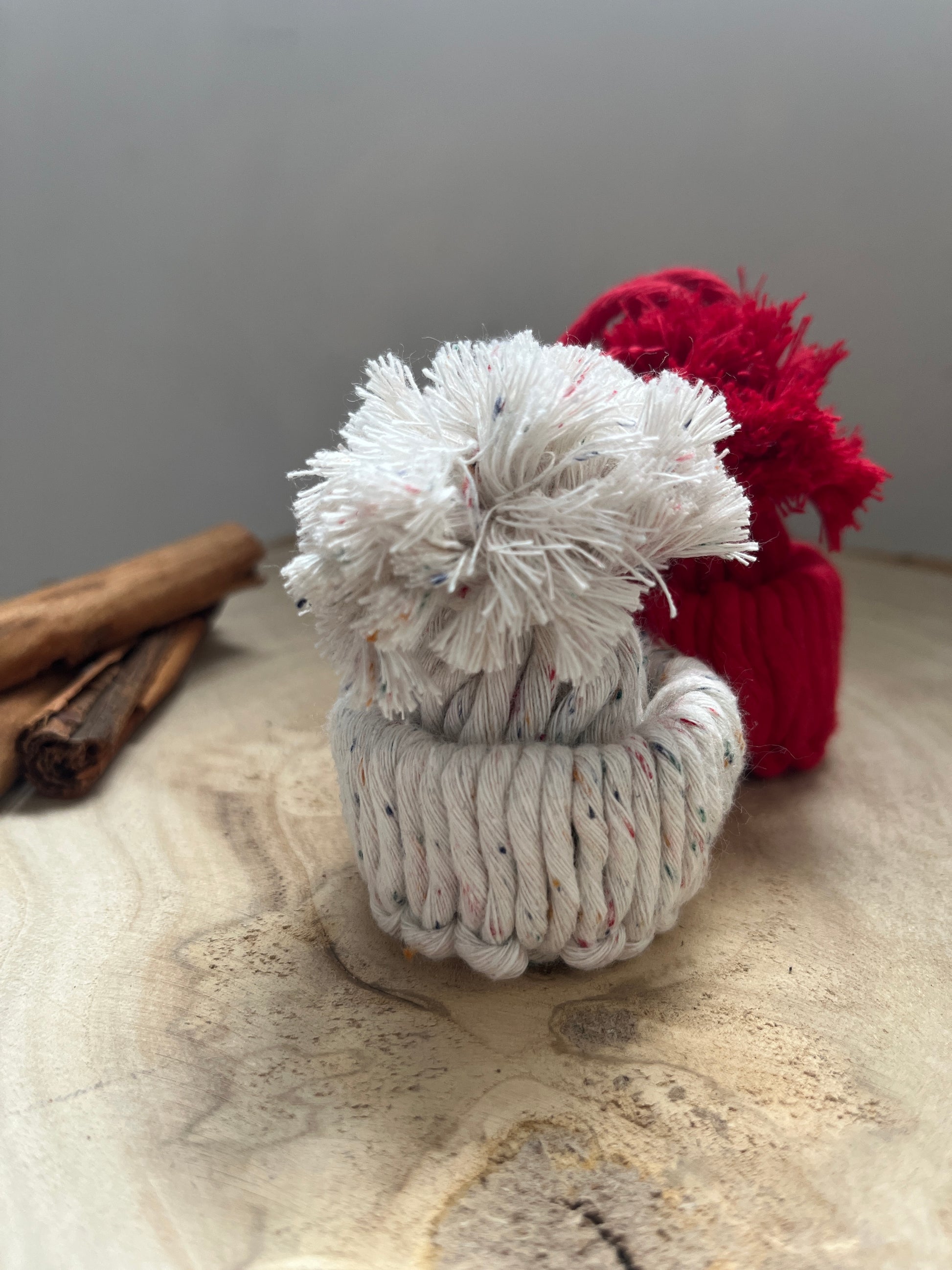 Two mini pom pom hat ornaments made with red and white yarns, on a wooden board with cinnamon sticks.