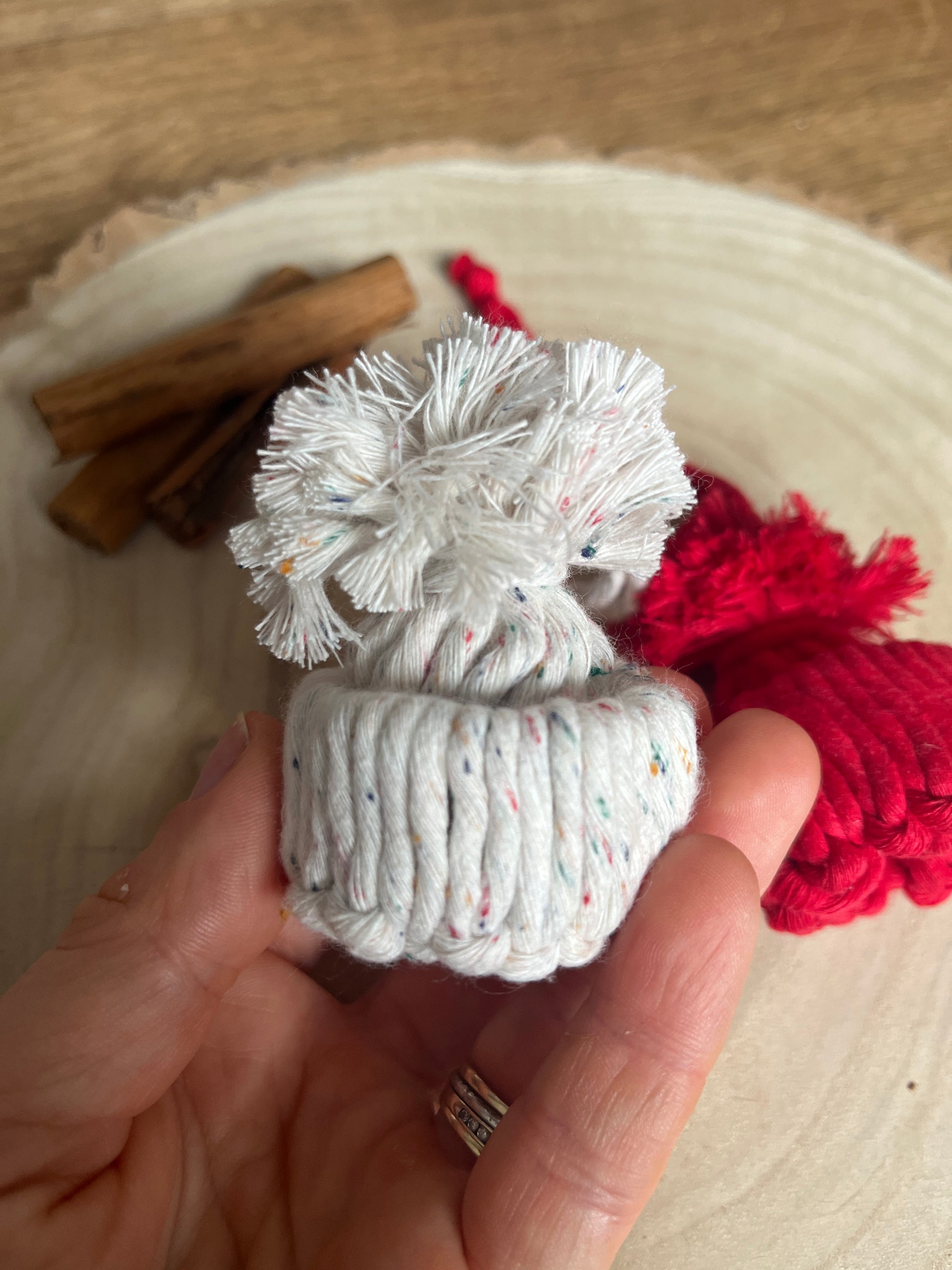 Hand holding a small white knotted pom pom hat ornament with a wooden surface in the background