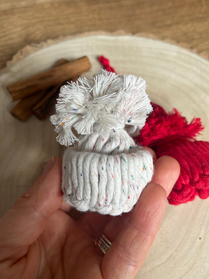 Hand holding a small white knotted pom pom hat ornament with a wooden surface in the background