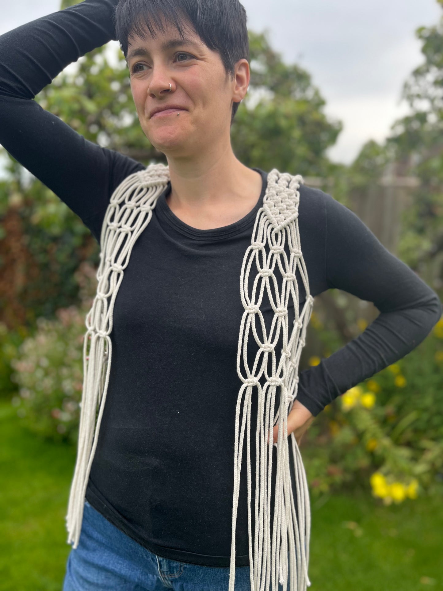 A person with short brown hair stands outside with a smile on her face, she is wearing a natural boho fringed waistcoat over a black long sleeved top, with a hand on the top of her head. 