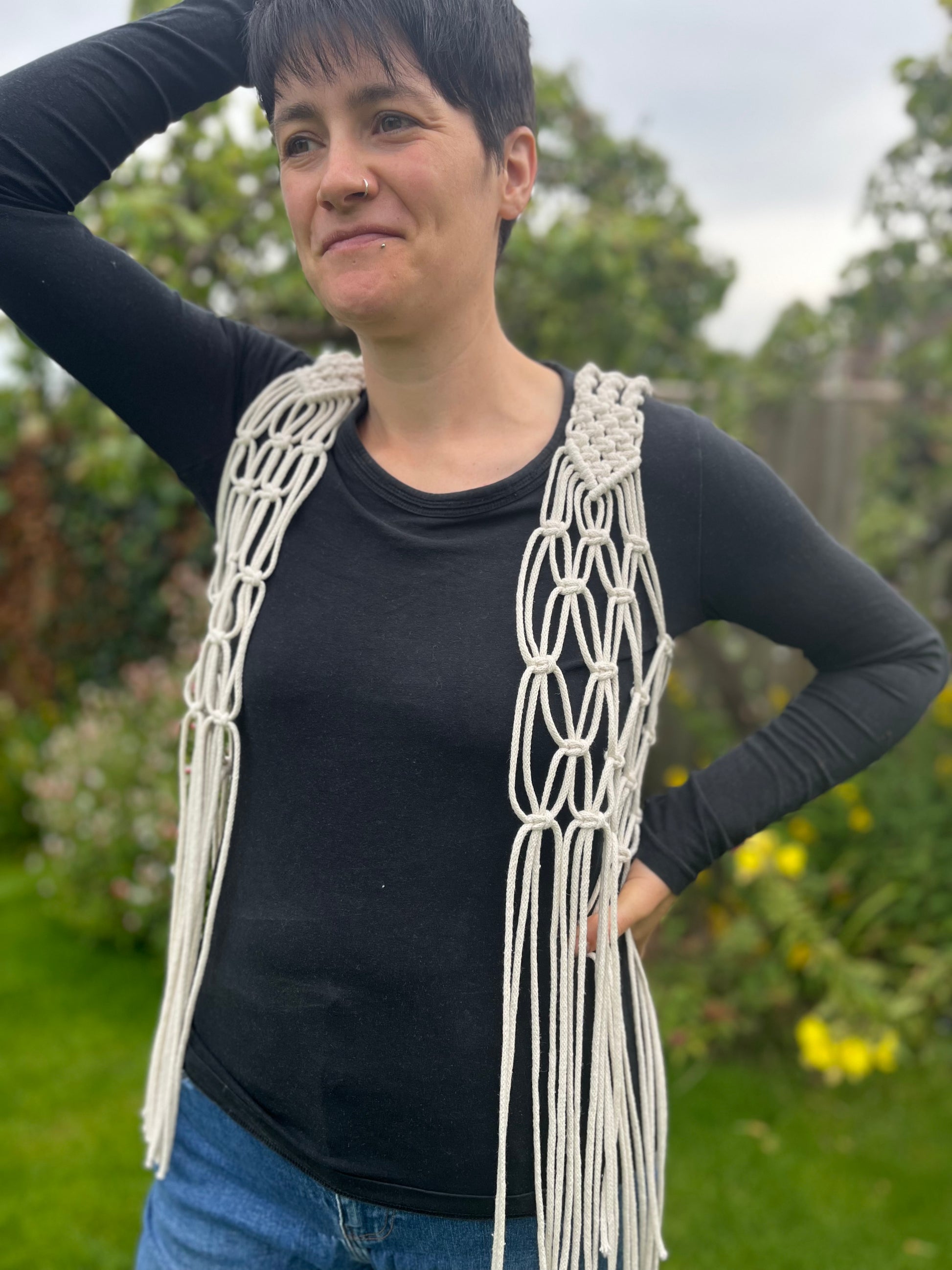 A person with short brown hair stands outside with a smile on her face, she is wearing a natural boho fringed waistcoat over a black long sleeved top, with a hand on the top of her head. 