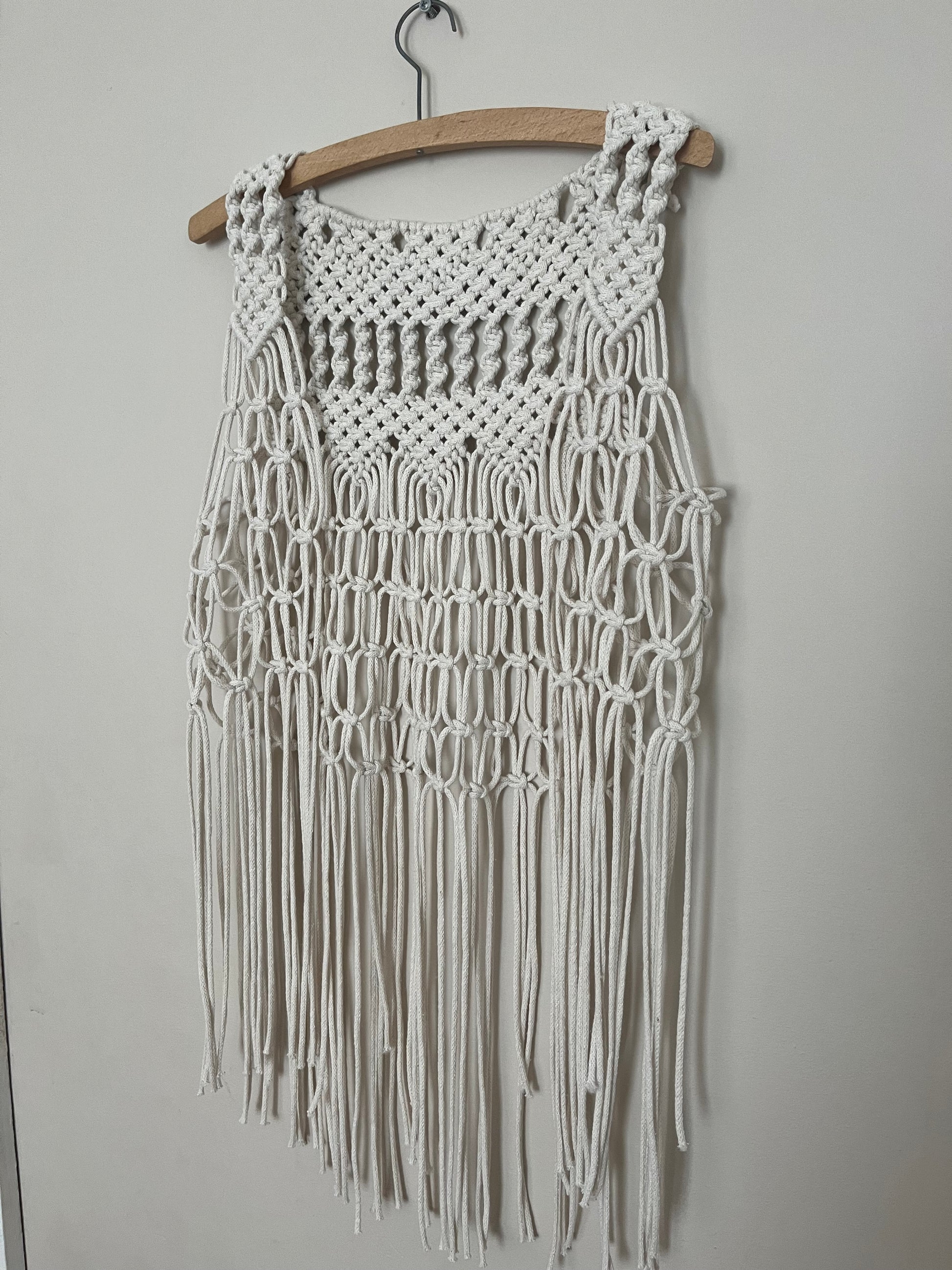 A handmade natural boho fringed waistcoat made with intricate macrame knots, hanging on a wooden coat hanger against a beige wall. 