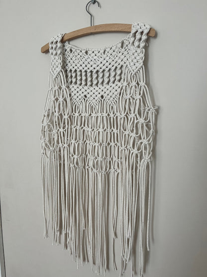 A handmade natural boho fringed waistcoat made with intricate macrame knots, hanging on a wooden coat hanger against a beige wall. 