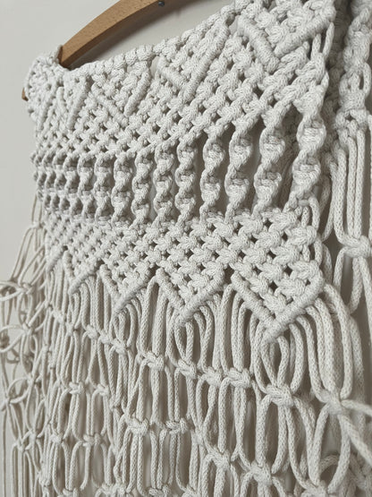 Close up of a handmade natural boho fringed waistcoat made with intricate macrame knots, hanging on a wooden coat hanger against a beige wall. 