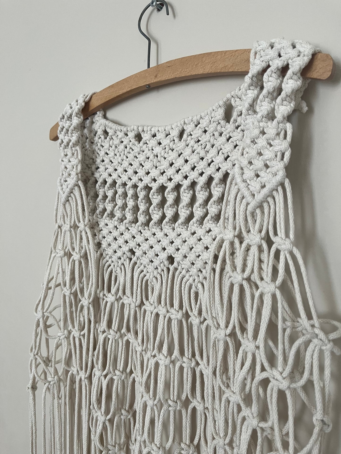 Close up of the intricate knot detail on a handmade natural boho fringed waistcoat hanging on a wooden coat hanger against a beige wall. 