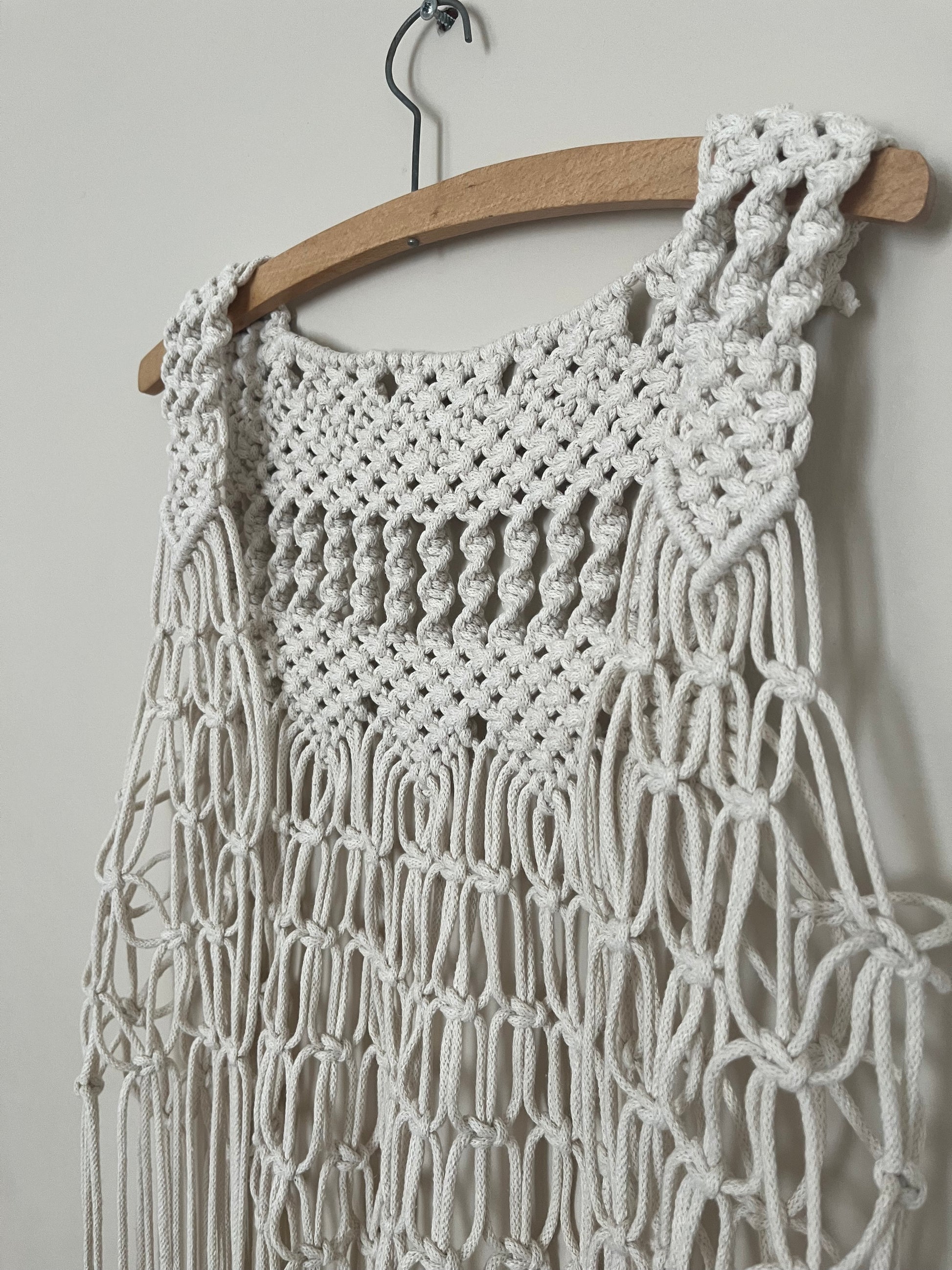 Close up of the intricate knot detail on a handmade natural boho fringed waistcoat hanging on a wooden coat hanger against a beige wall. 